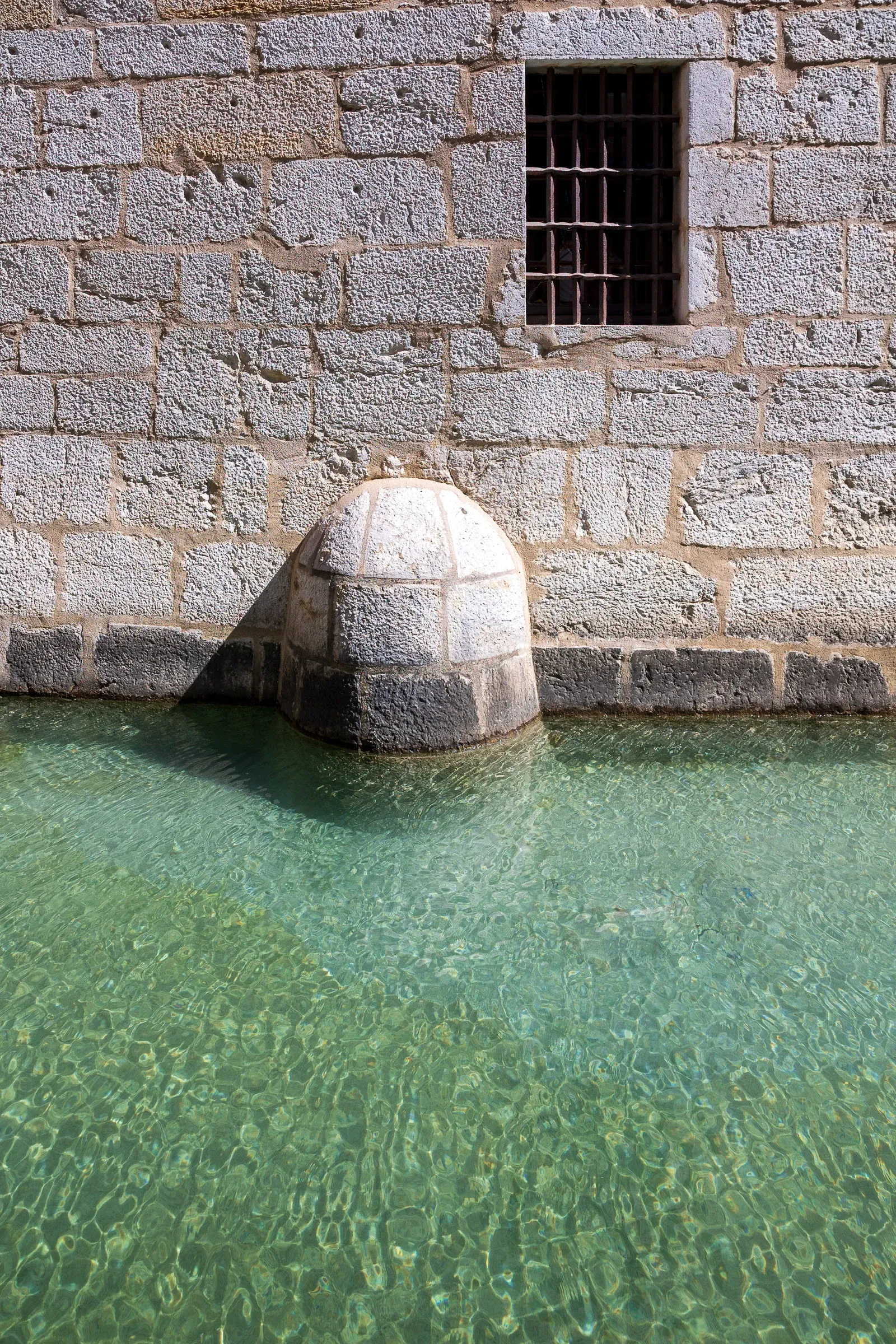 Stone bridge pillar rising from crystal-clear turquoise canal water beside a barred window, Annecy old town