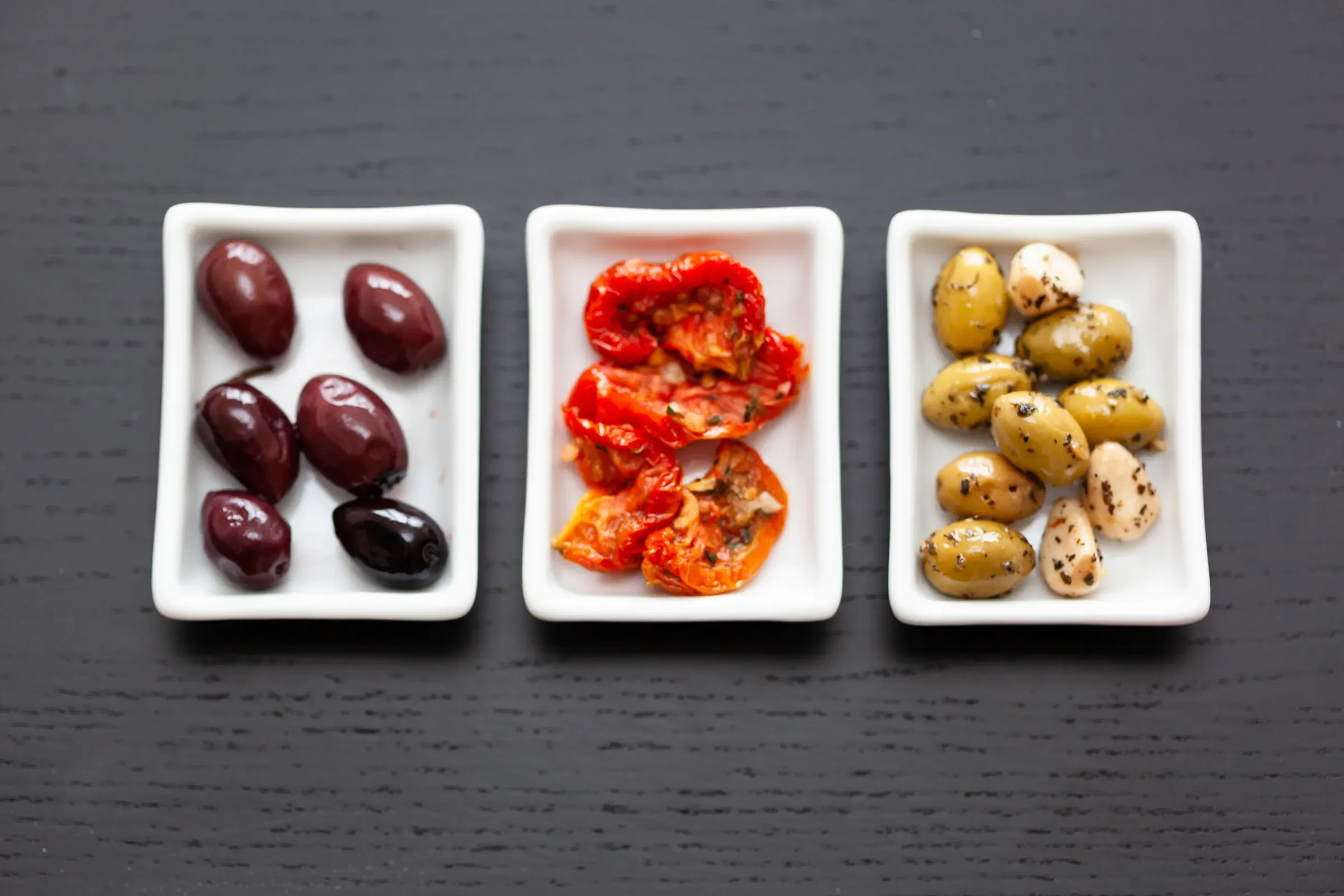 Three white ceramic dishes on dark slate — Kalamata olives on the left, confit tomatoes in the centre, herb-marinated green olives and garlic on the right
