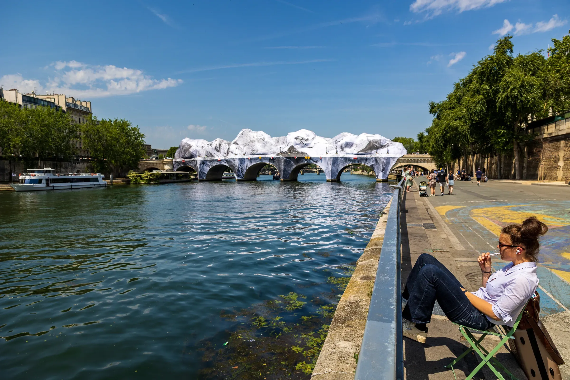 Woman seated on a folding chair at the quai, the Pont Neuf wrapped in JR's trompe-l'œil cave installation across the Seine, a tour boat passing, Paris