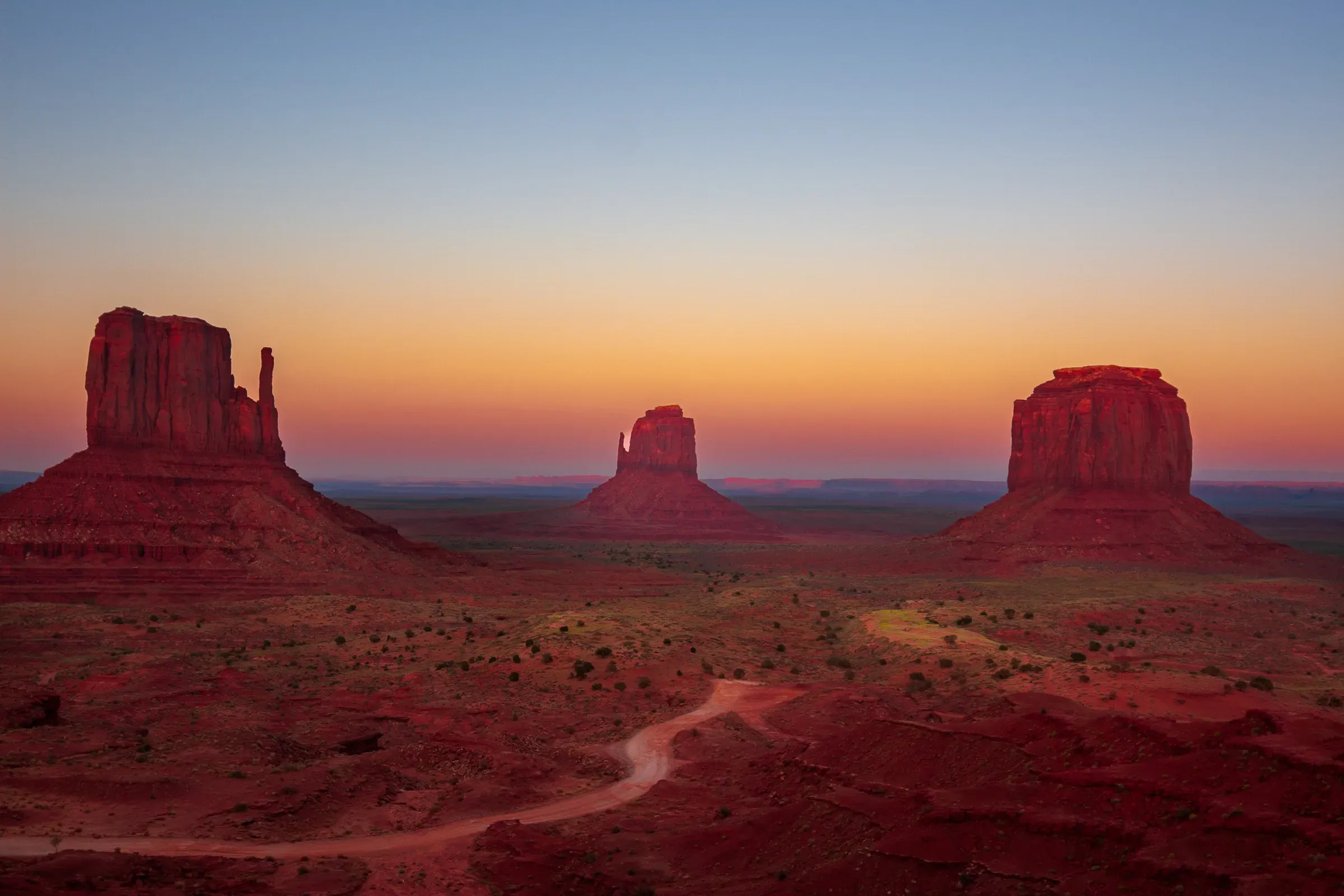 Three sandstone buttes at dusk, Monument Valley — orange sky fading to deep blue, dirt road winding through the red desert floor