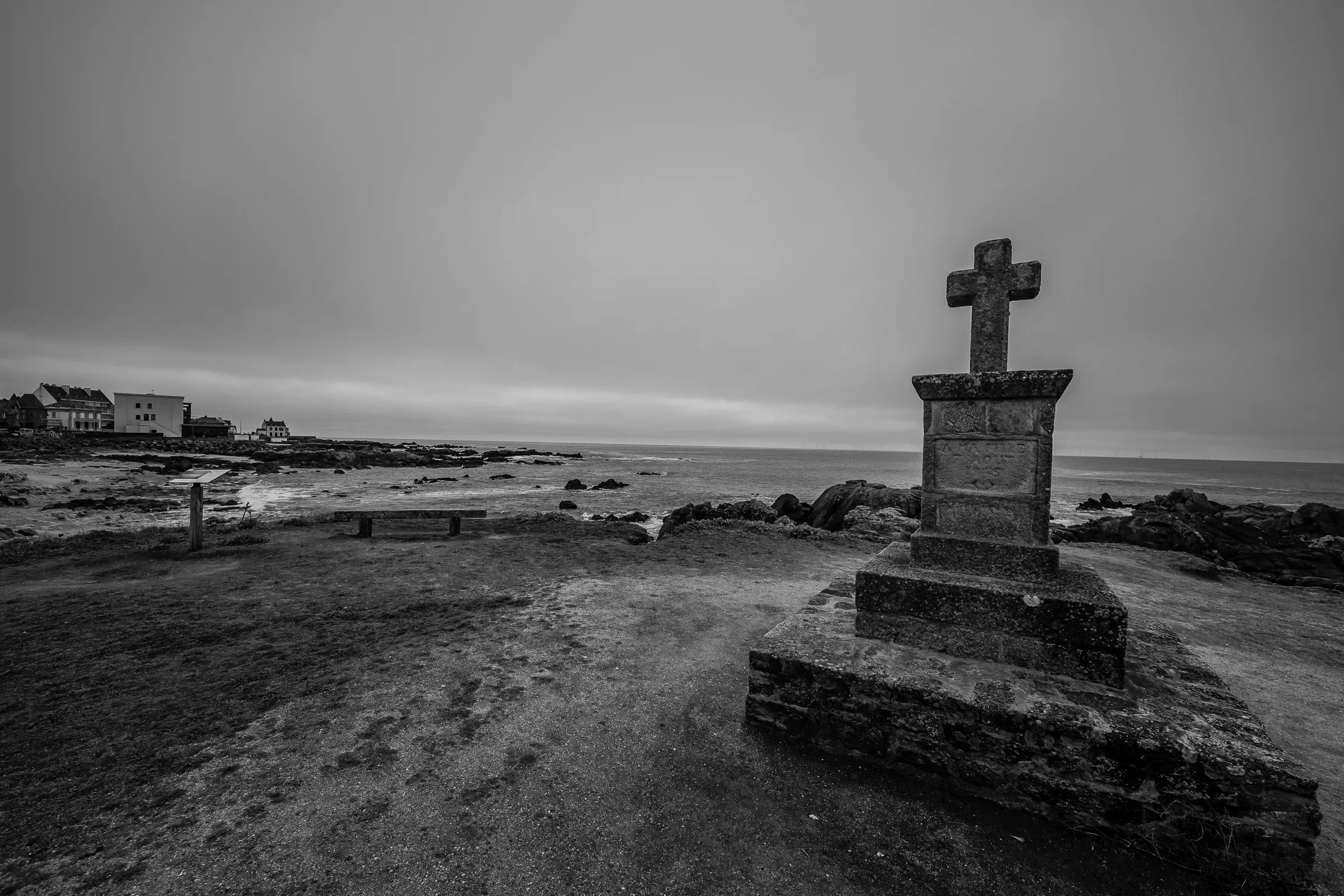 Black and white — a stone calvary cross on a coastal headland, overcast sky, rough sea and houses behind
