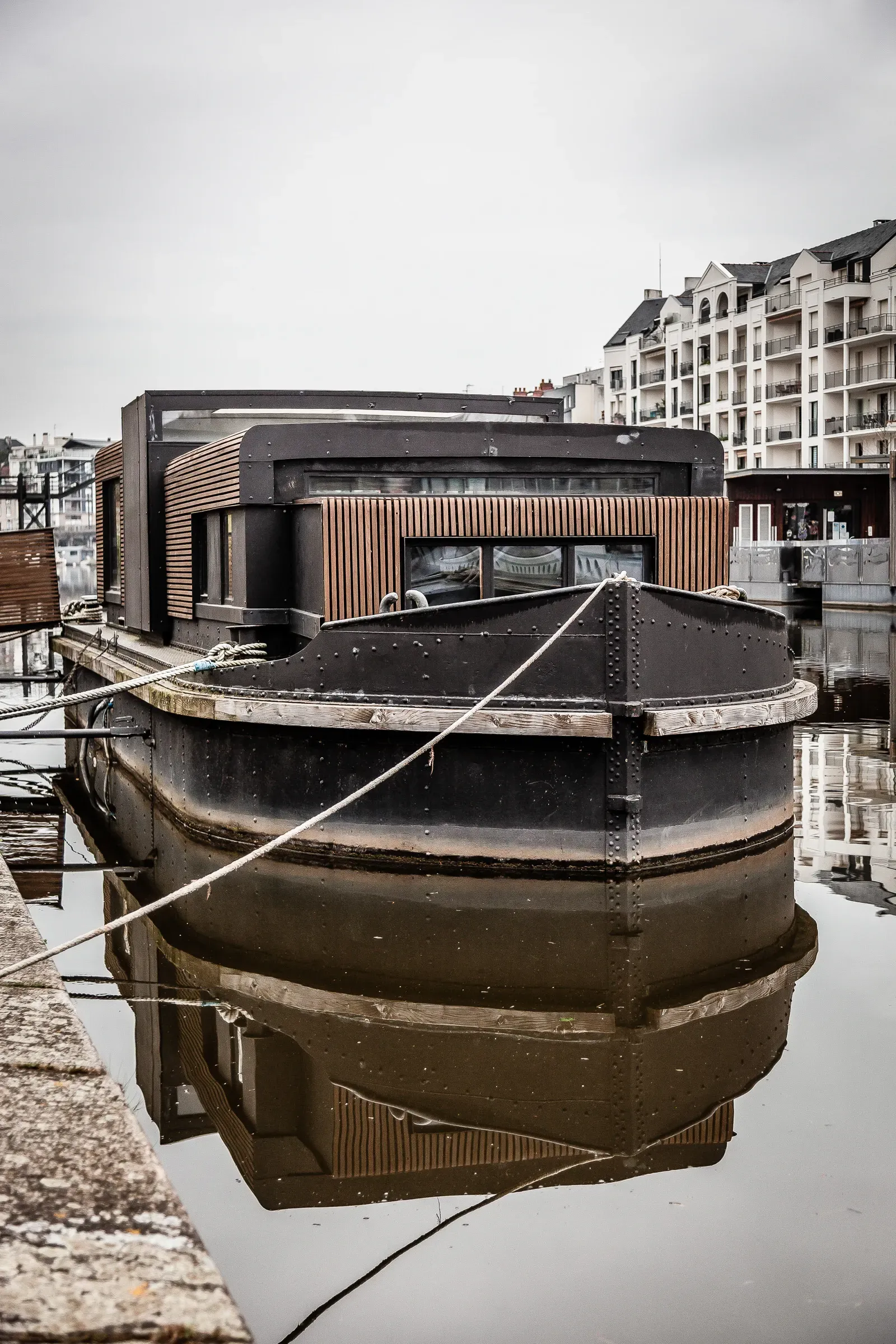 A sleek dark houseboat with timber cladding, moored in an urban harbor, modern apartment blocks behind, overcast sky