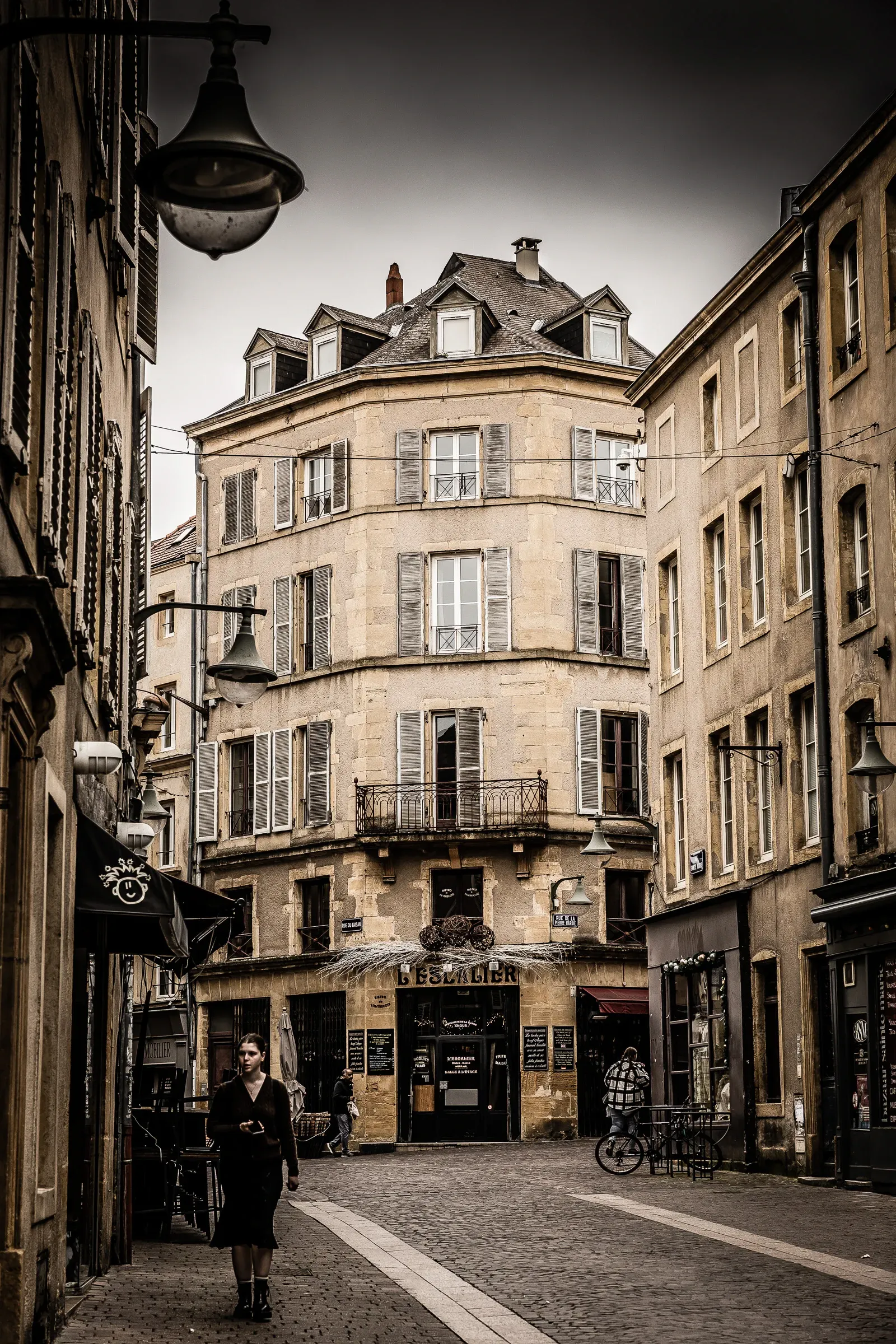 A woman walking away down a narrow cobblestone street lined with honey-stone buildings, a street lamp overhead, Metz