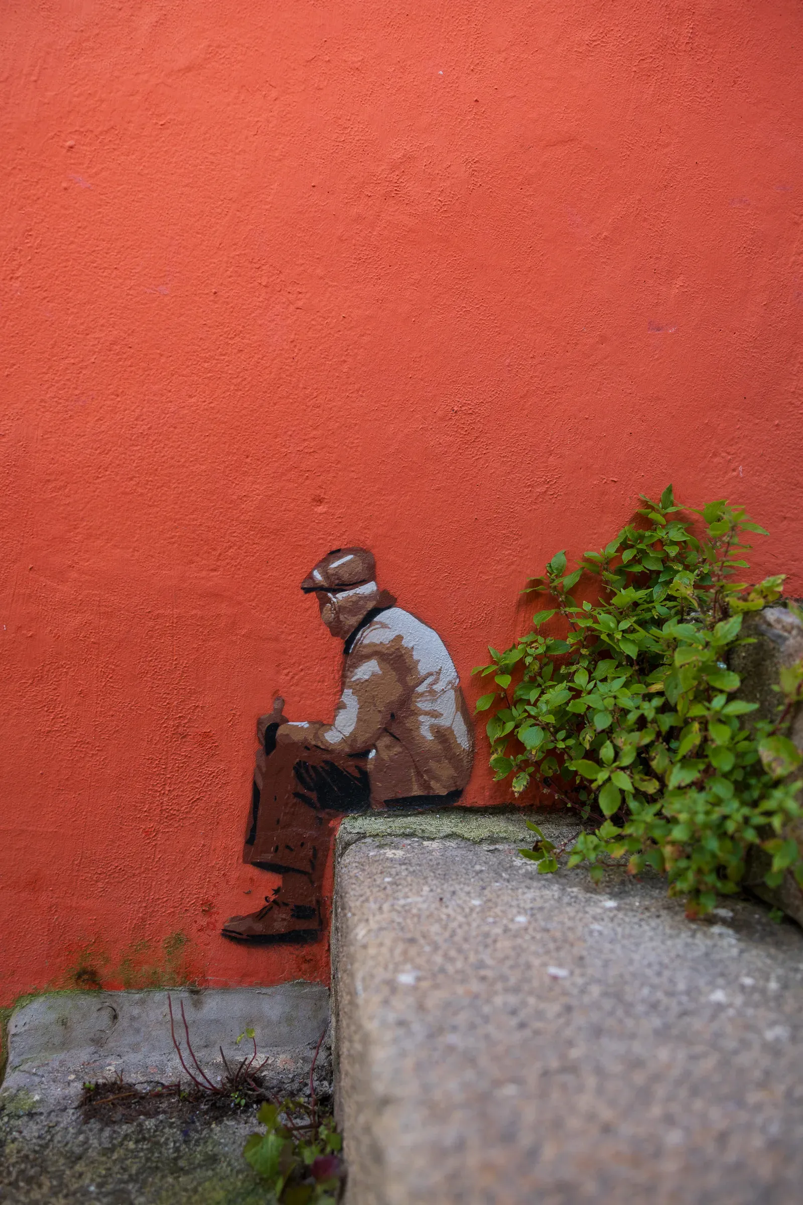 A small painted figure of a man sitting on stone steps against a deep orange-red wall, green plants beside him