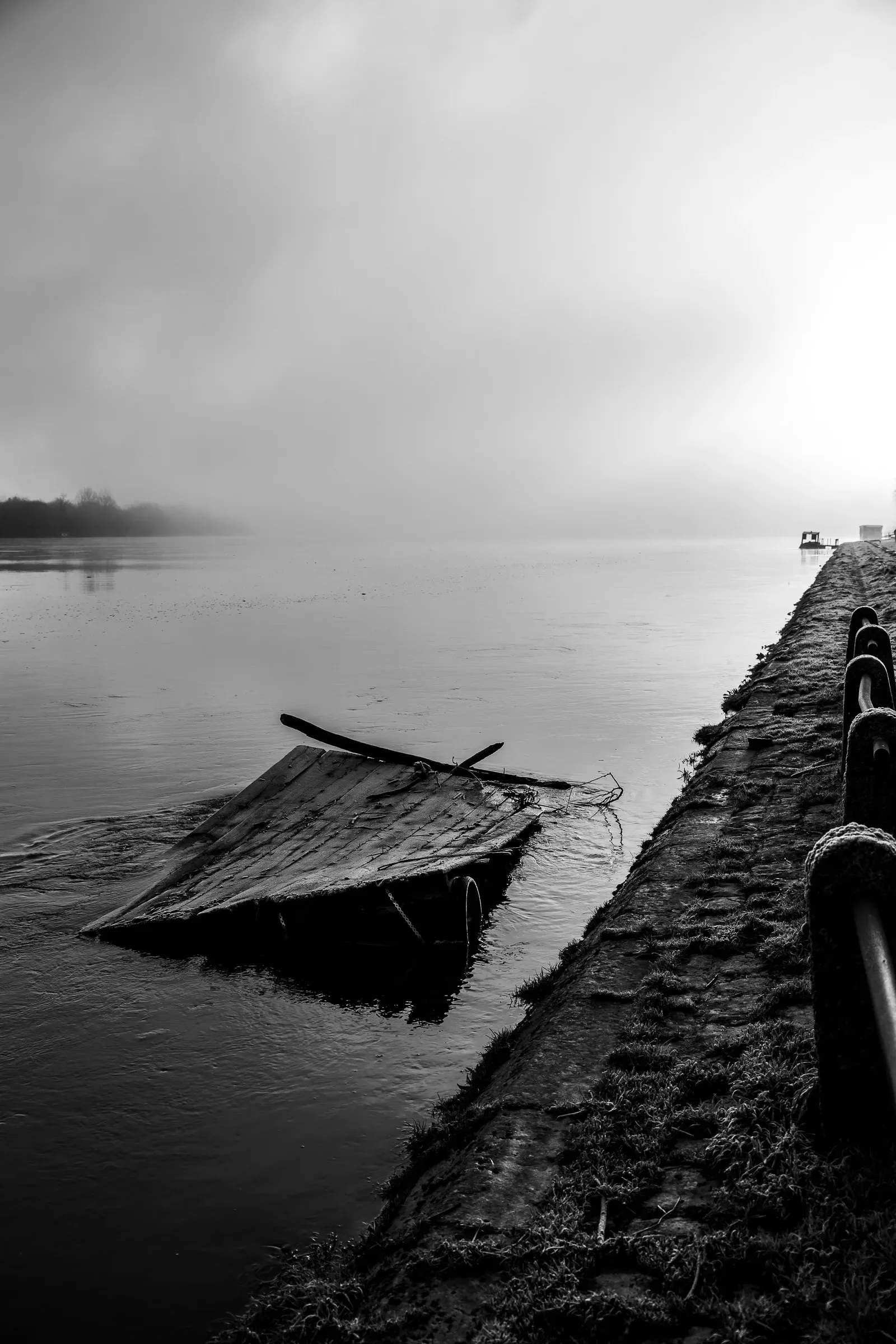Black and white — overturned wooden boat half-submerged at the edge of a stone quay, fog-blanketed river stretching behind, Saumur
