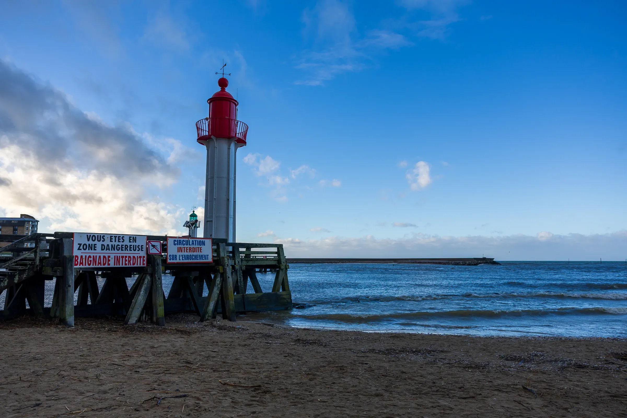 Red and white Trouville lighthouse beside a wooden pier with Baignade Interdite warning signs, calm blue sea and patchy clouds, Trouville