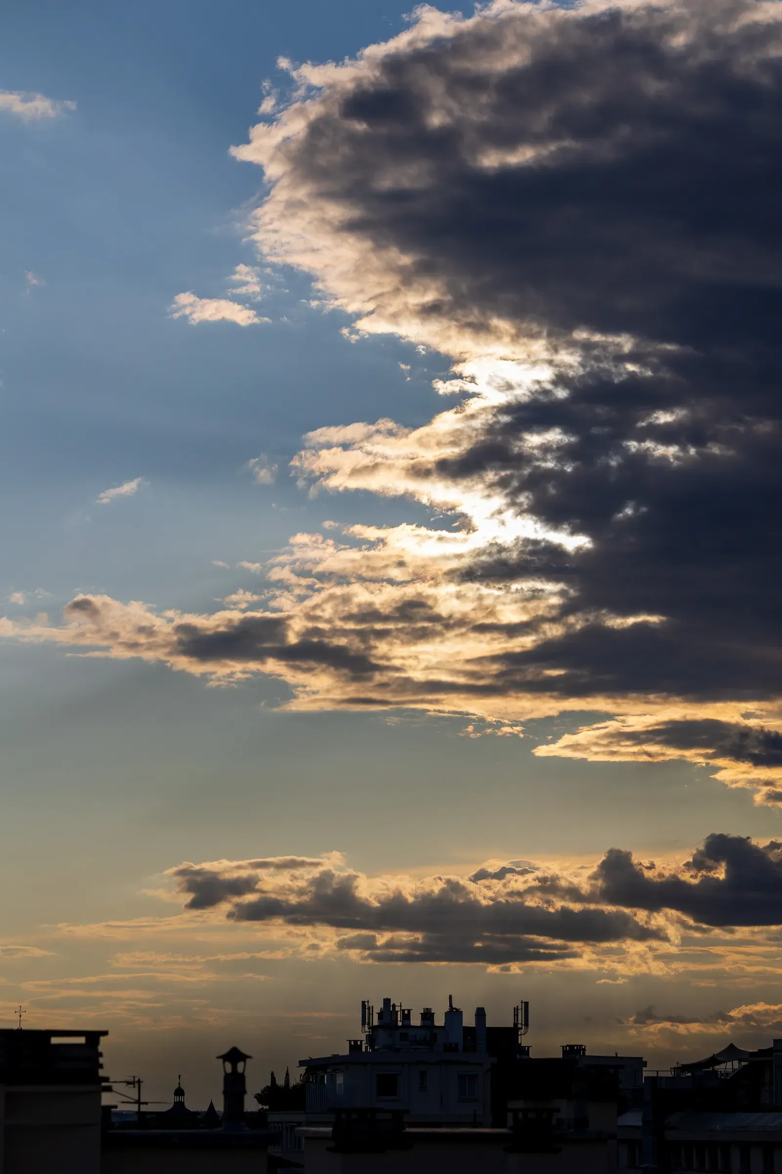 Dramatic sunset sky above Toulouse rooftops — layered clouds backlit in gold and dark blue, minaret and building silhouettes along the bottom, Toulouse