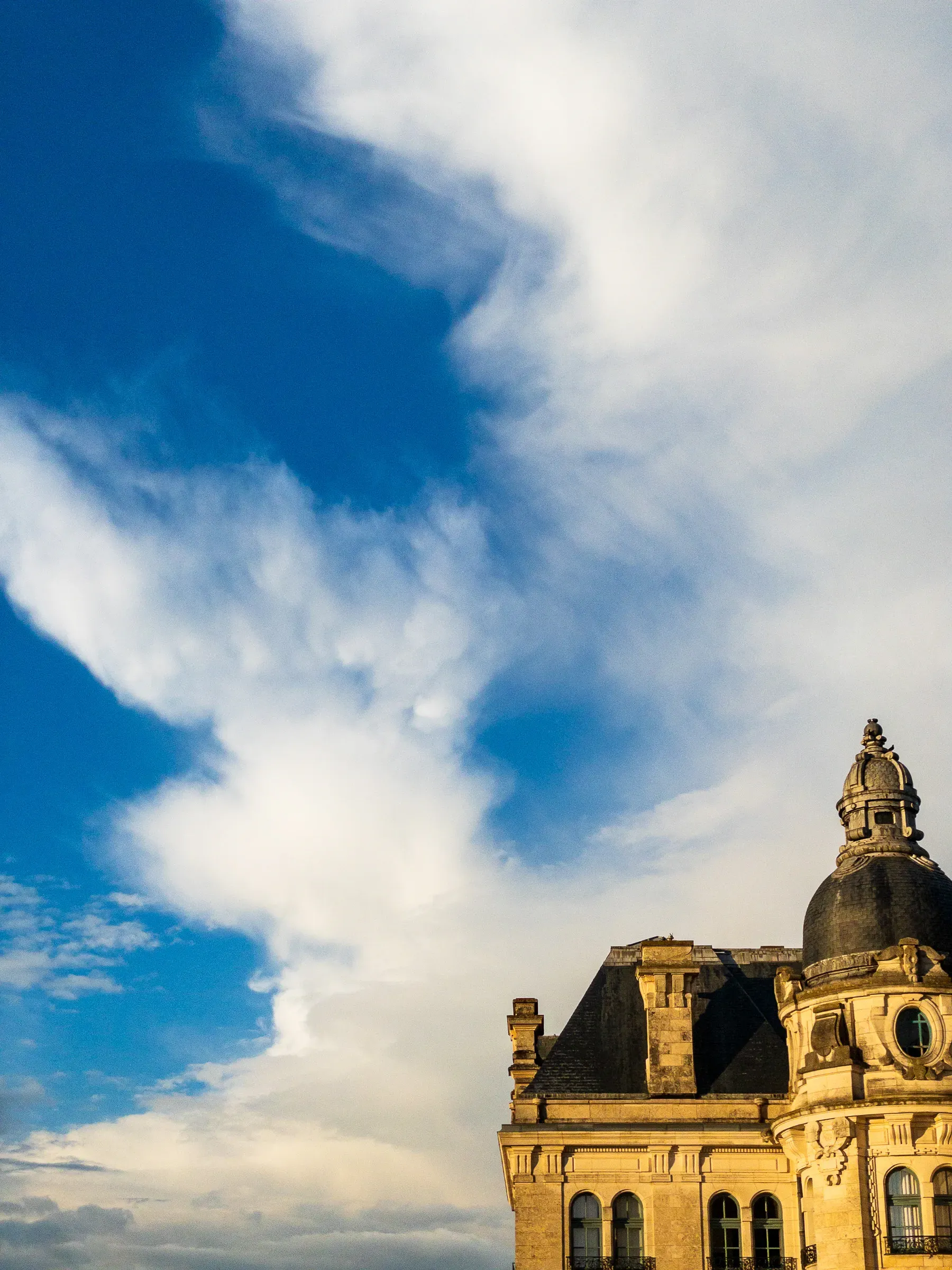Corner of a classical stone building with an ornate copper dome, warm golden light on the facade, dramatic blue sky filled with wispy white clouds