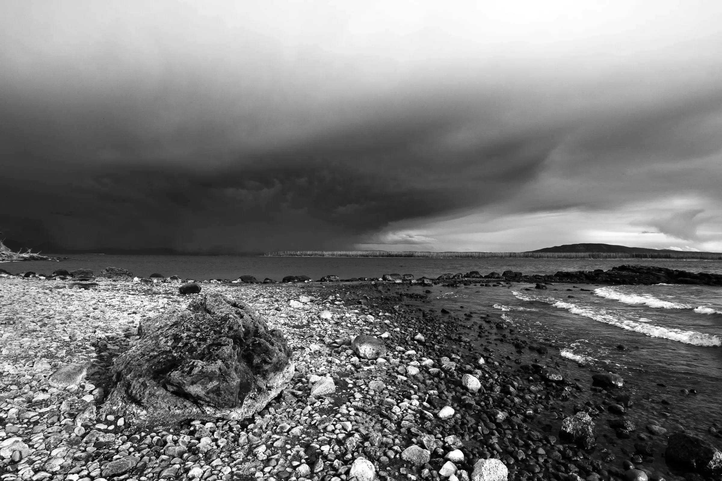 Rocky shingle beach with a large moss-covered boulder in the foreground, stormy clouds rolling in over a grey sea, a distant headland on the horizon