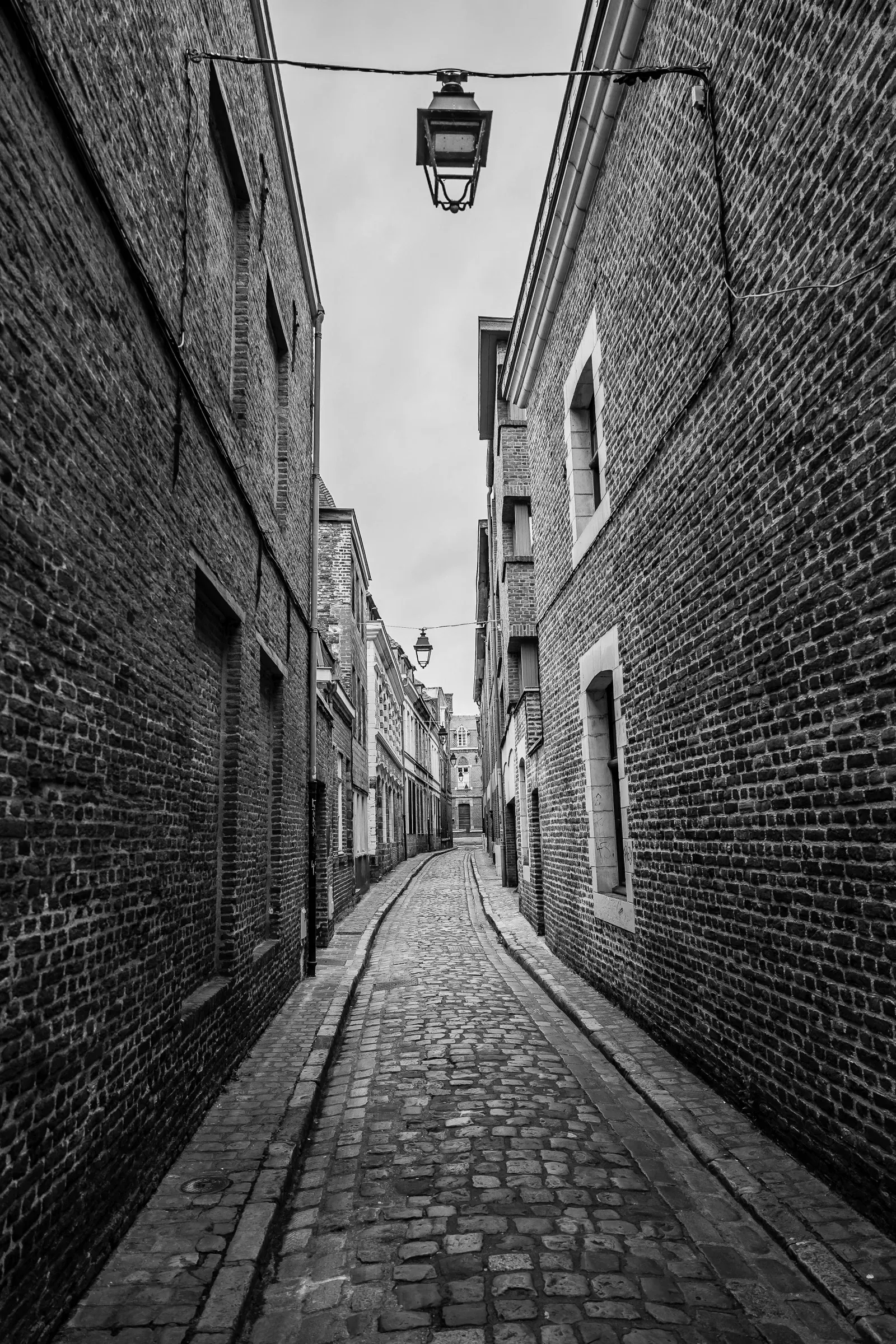Narrow cobblestone alley between brick buildings with a hanging street lamp, black and white, Lille