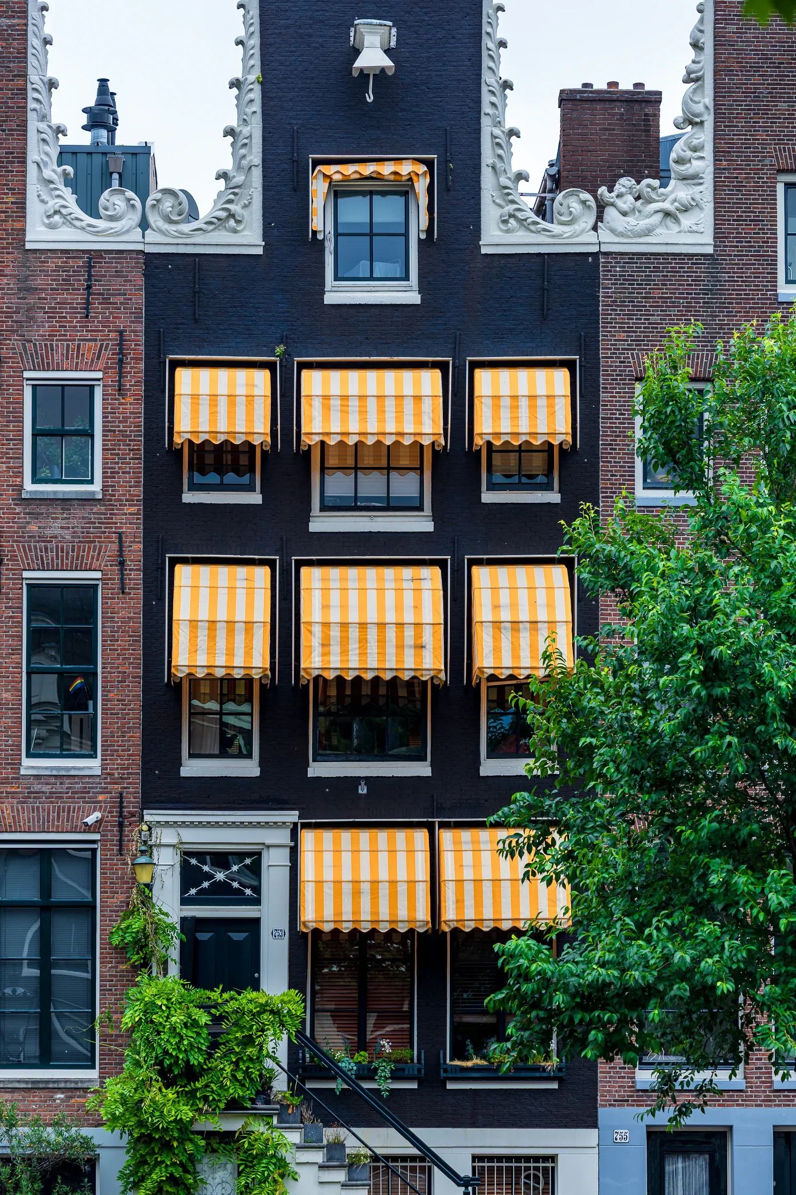Dark narrow canal house facade with an ornate white carved gable, yellow-and-white striped awnings on every window