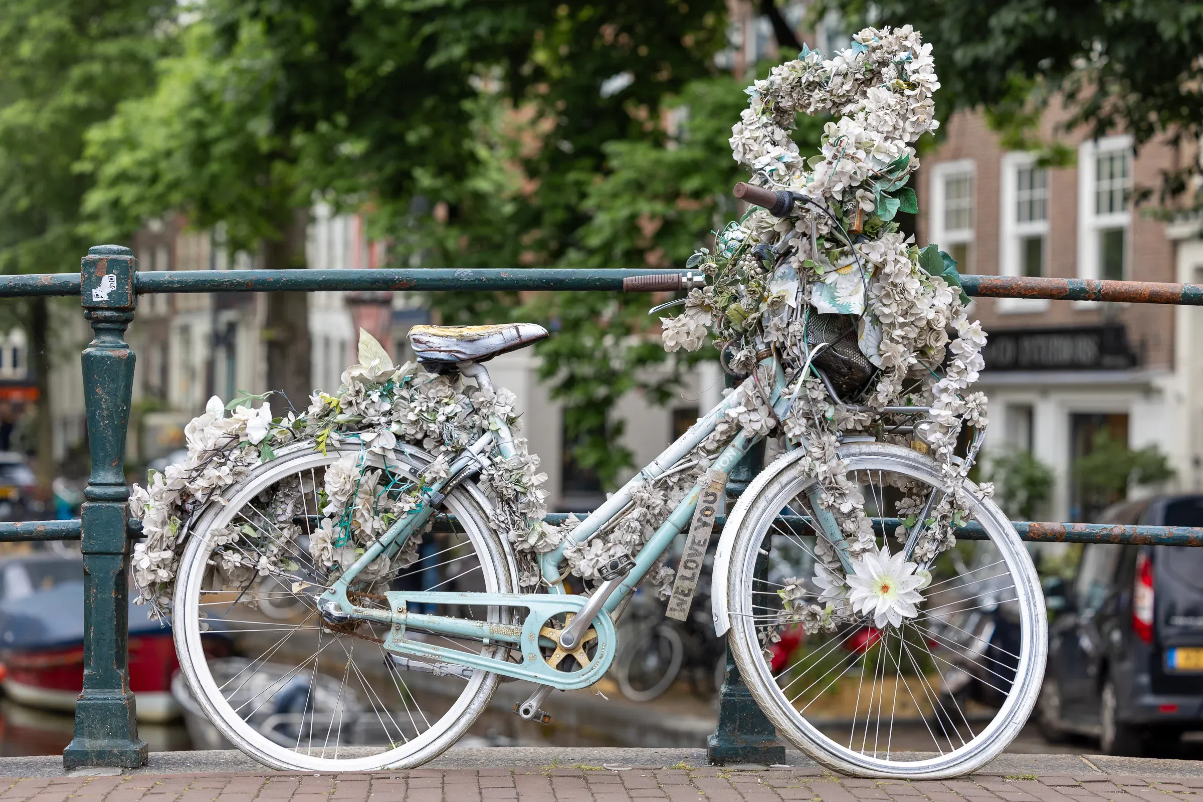 A sky-blue bicycle smothered in white fabric flowers, chained to a canal bridge railing, Amsterdam canal houses behind
