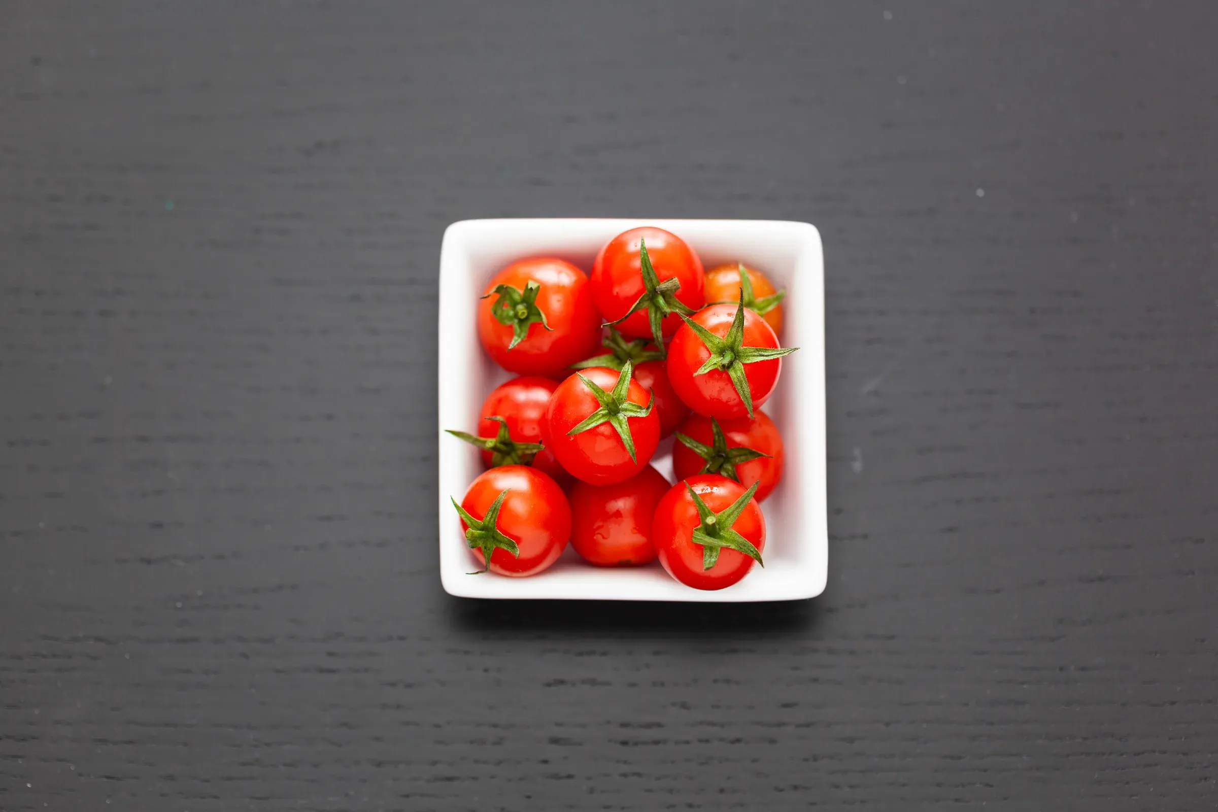 Cherry tomatoes with stems in a white square ceramic dish, overhead, dark slate surface
