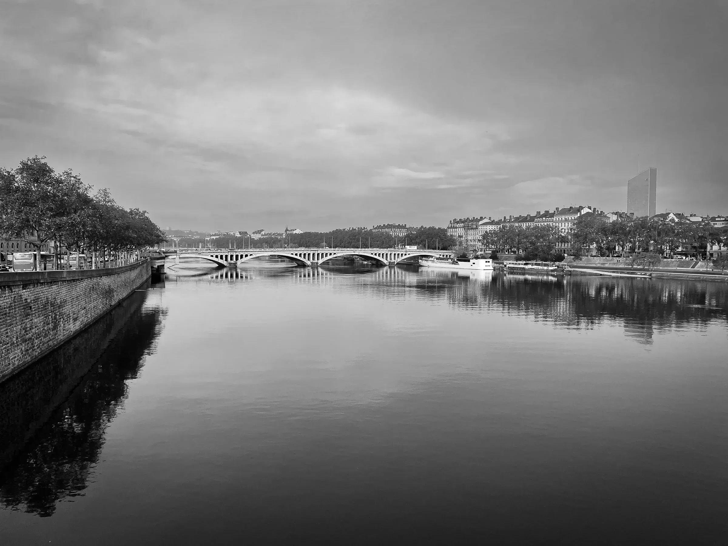 Black and white view of the Rhône river with a stone bridge and modern tower reflected in calm water, Lyon