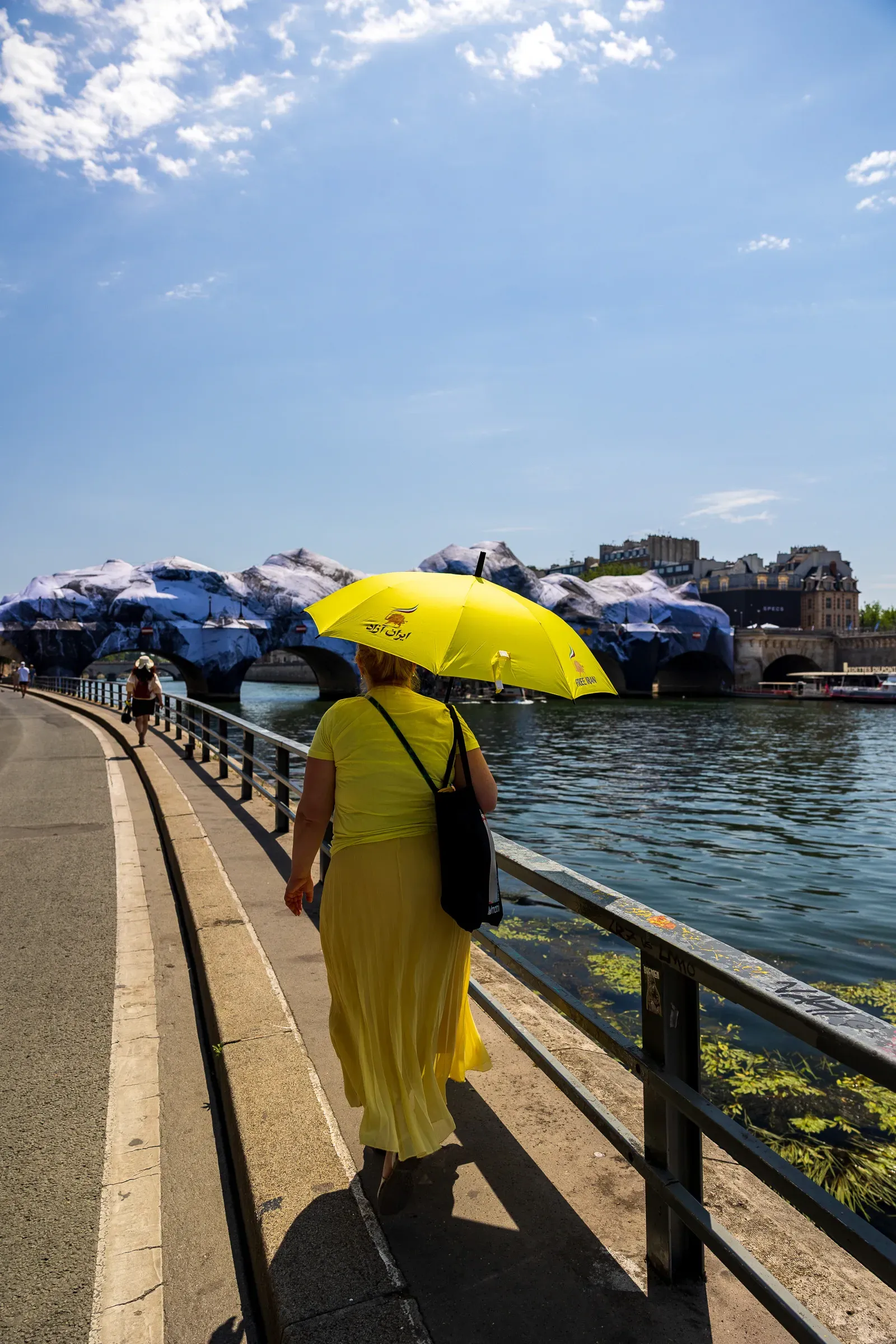 Woman in a yellow dress carrying a yellow umbrella, walking the riverside path with JR's caved Pont Neuf installation across the water, Paris