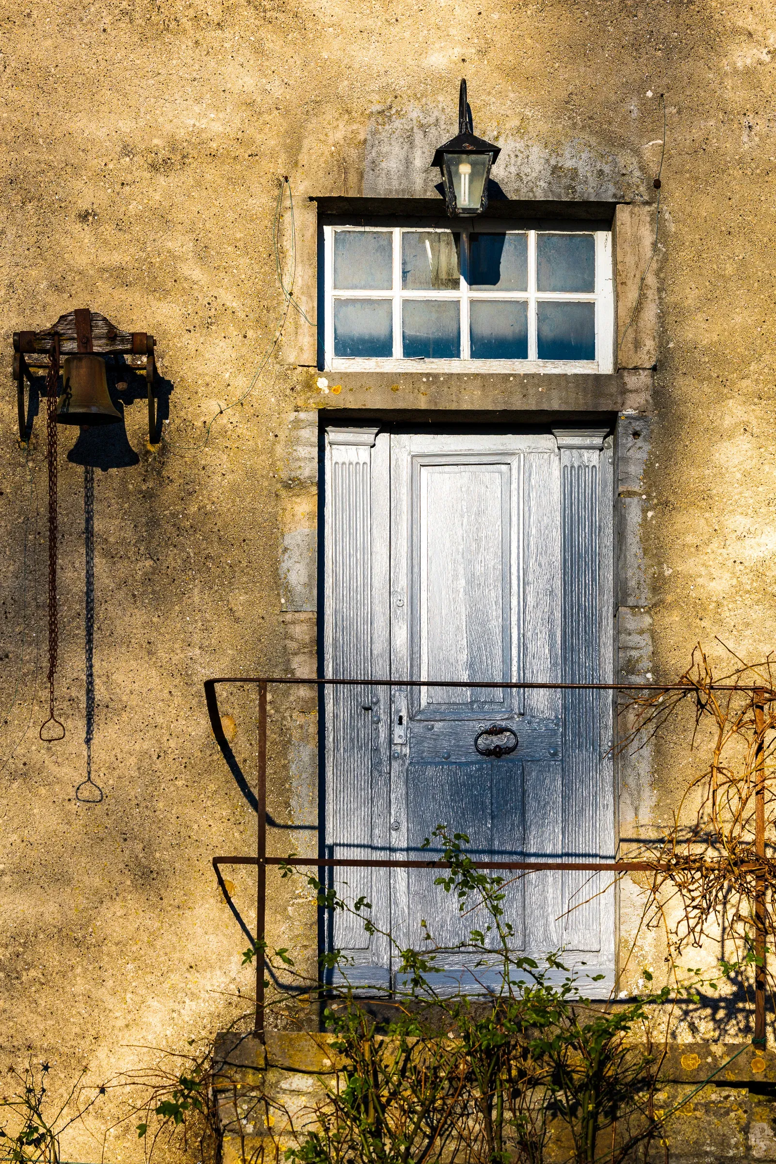 Weathered stone farmhouse facade with a pale blue double door, iron lantern above, old bell on the left, bare climbing vine