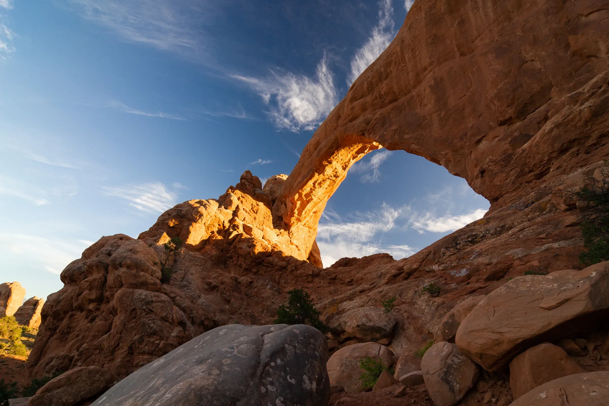 Natural sandstone arch from below, Arches National Park — warm afternoon light on the rock face, blue sky and cirrus clouds through the opening
