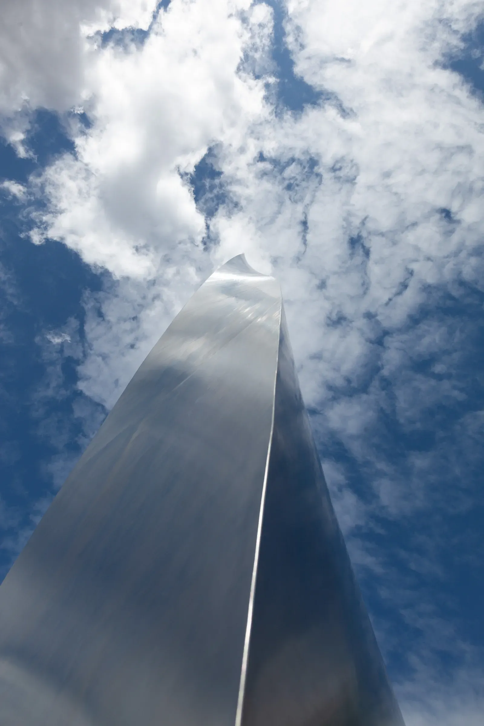 Looking straight up at a polished steel spire, its mirrored surface reflecting a cloud-scattered blue sky
