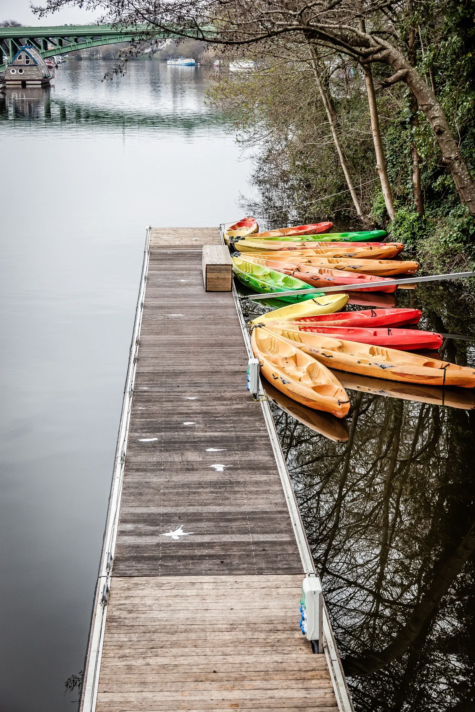 A row of colorful kayaks lined up on a wooden dock along a grey river, a green metal bridge in the background