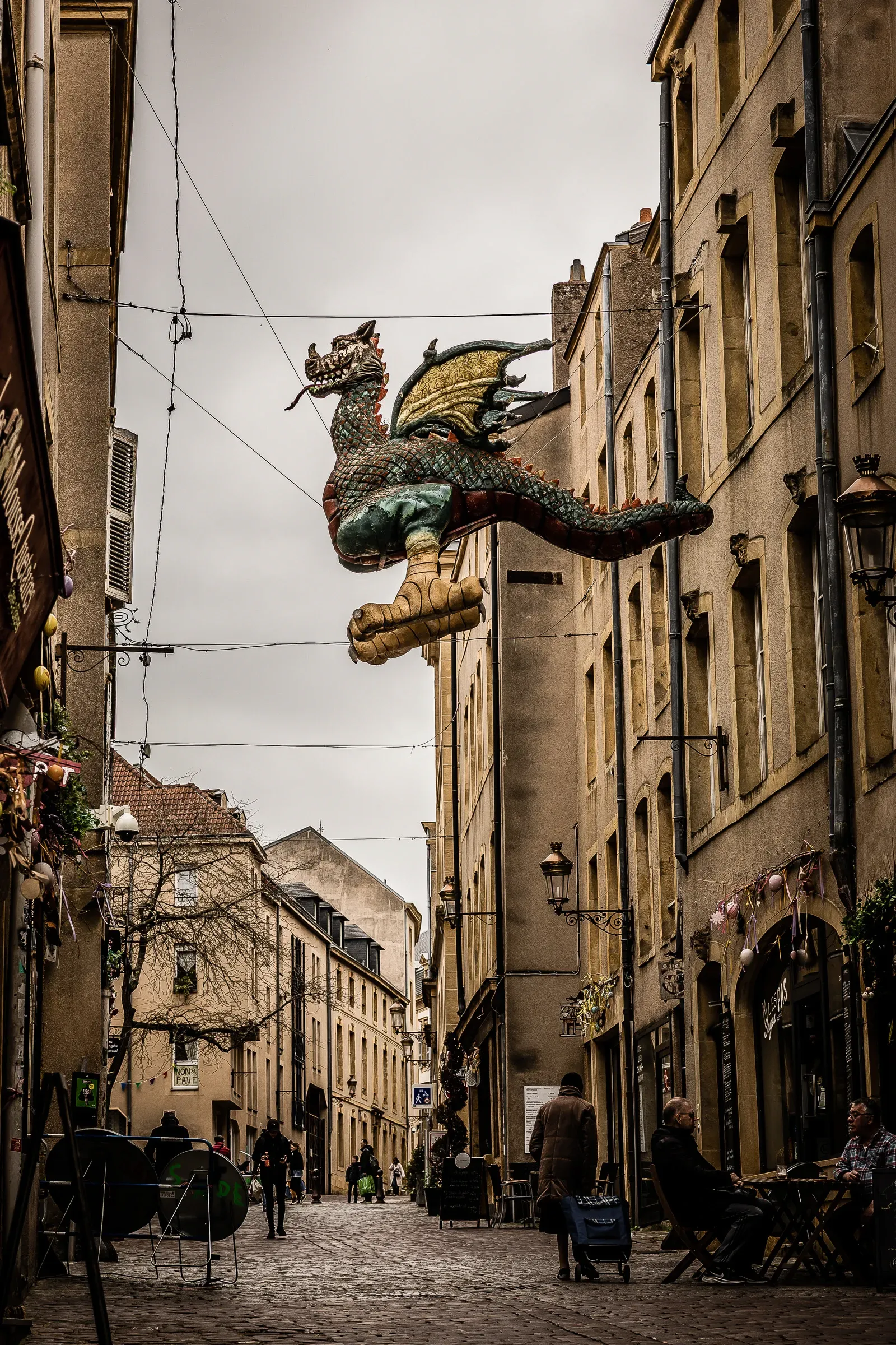 The Graoully dragon sculpture suspended in mid-air across a narrow old street between honey-stone facades, diners below