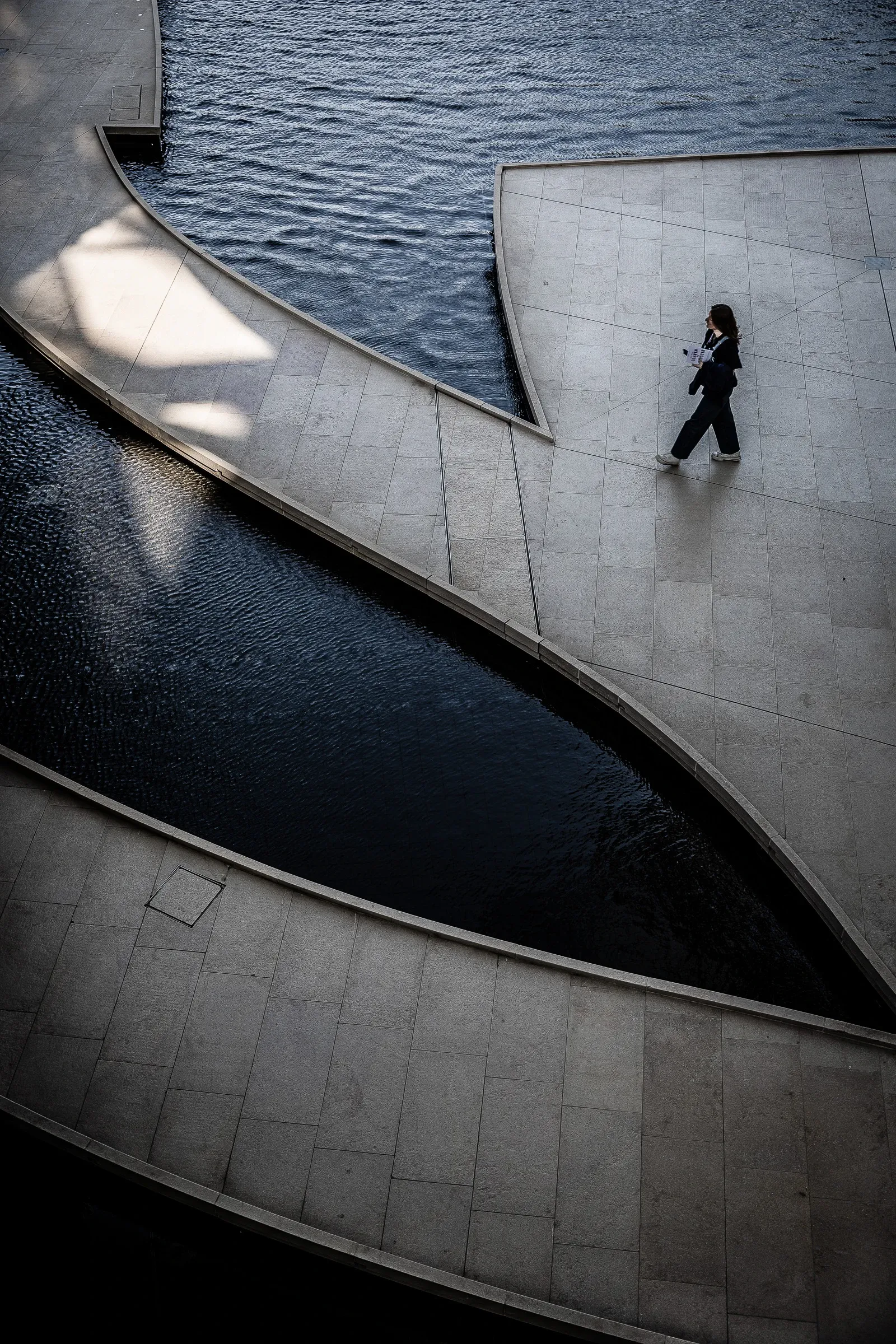 Aerial view of a person walking across a wave-patterned stone plaza beside dark water channels
