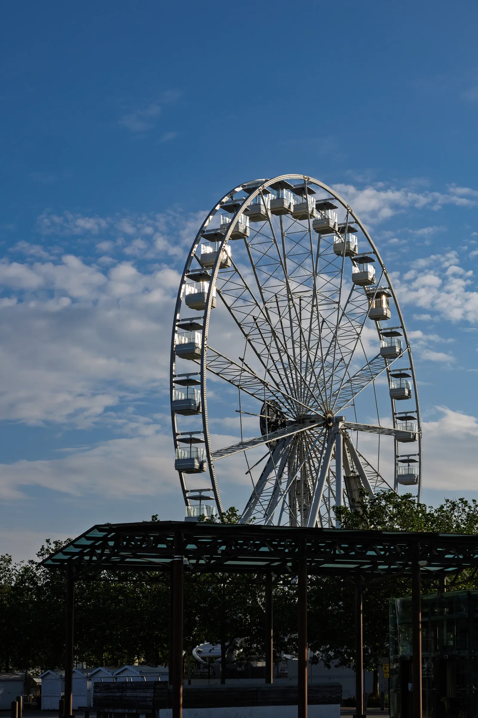 White Ferris wheel against a blue sky with light clouds, a glass-roofed canopy structure in the foreground, trees behind