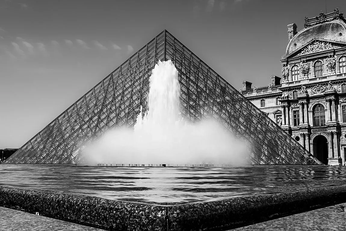 Louvre glass pyramid with a fountain spray rising in front of it, the classical stone facade of the Richelieu wing filling the background