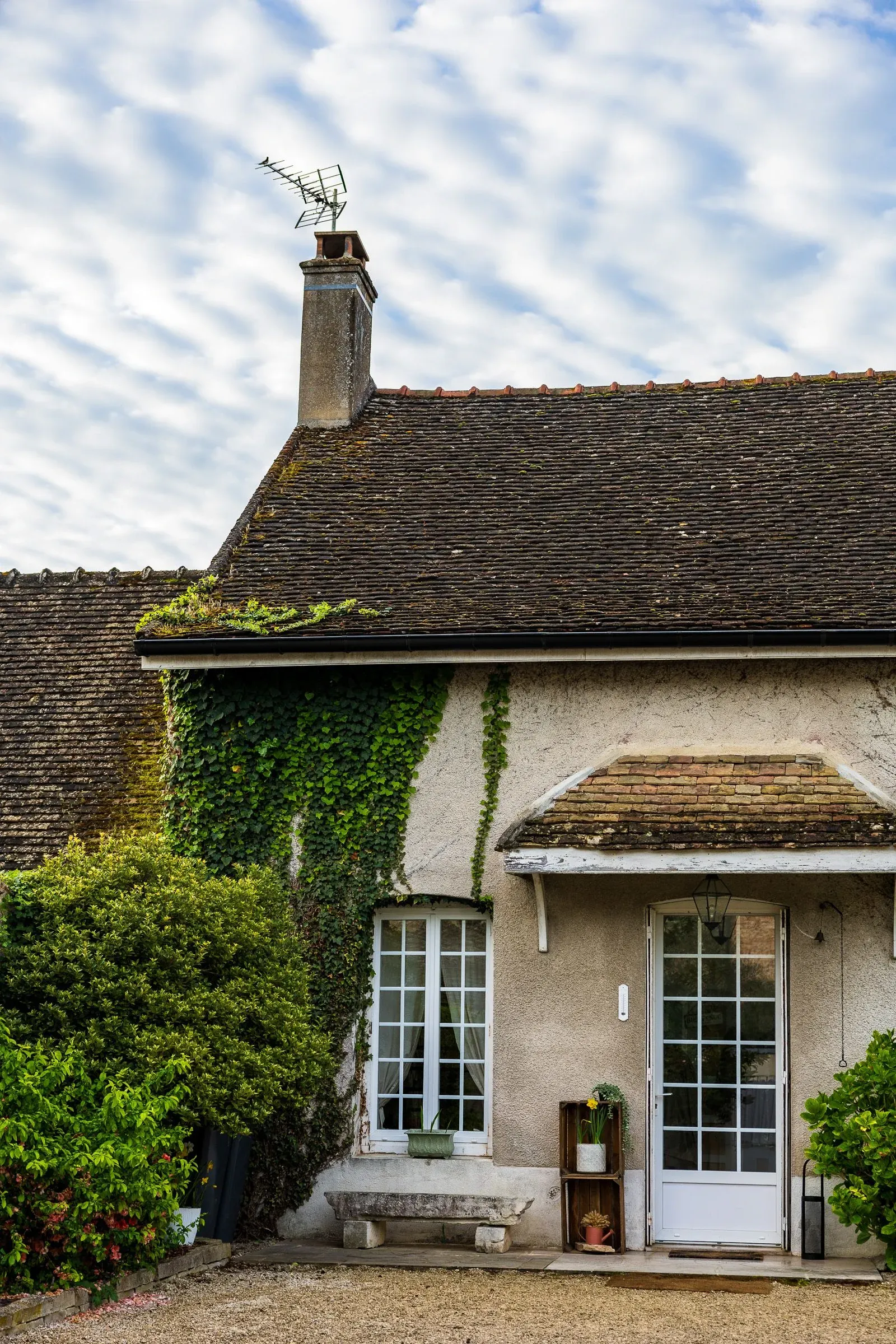 Gable end of an old tiled-roof house with an ivy-covered wall, a chimney topped with a TV aerial, dappled clouds above