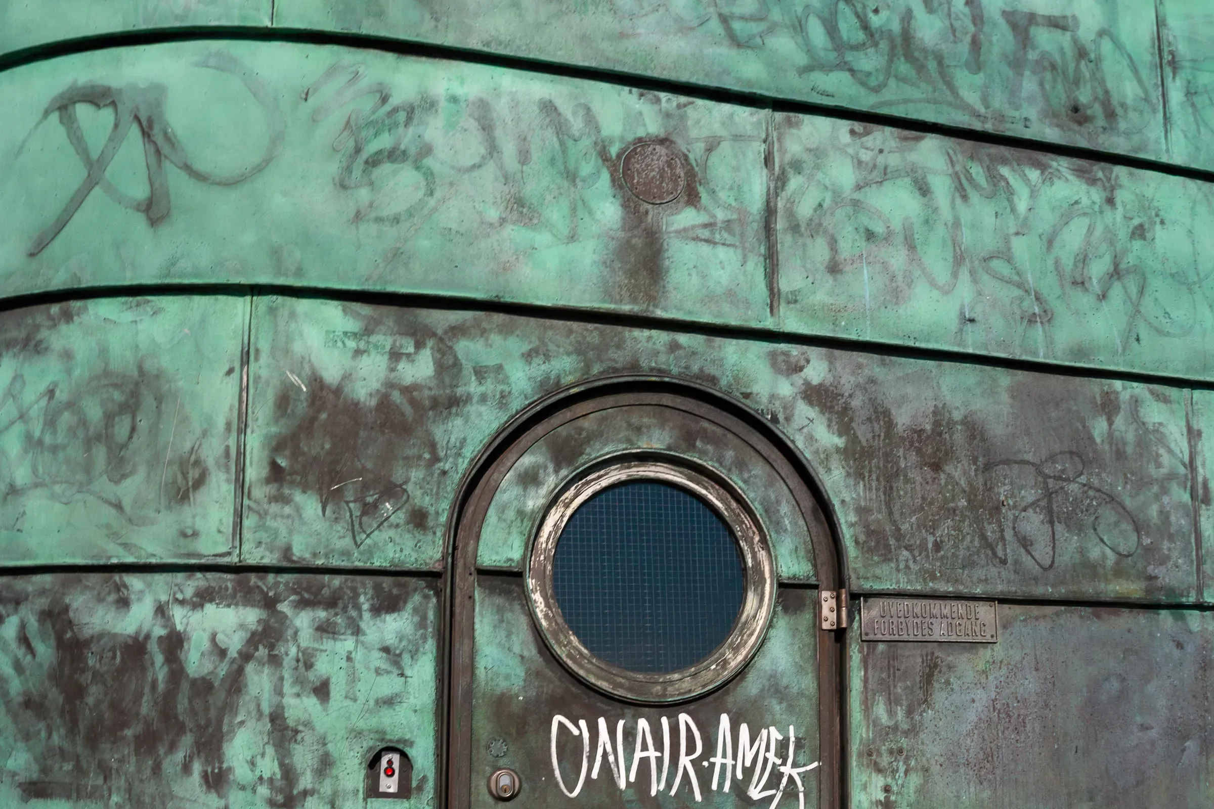 Green oxidized copper surface covered in graffiti with a round porthole door