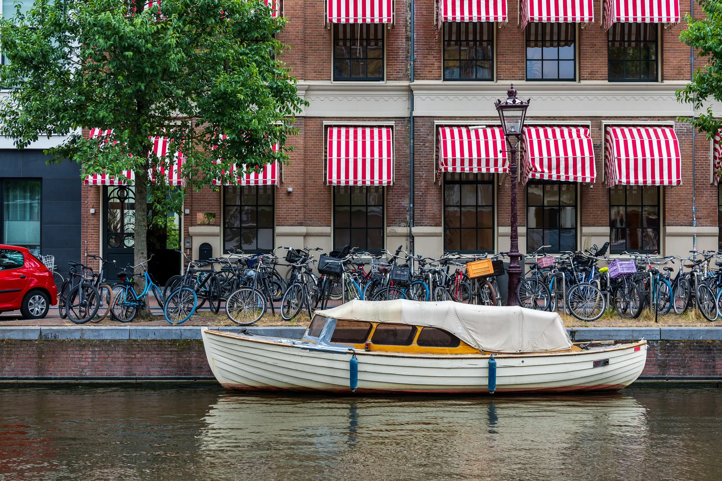 White wooden boat moored on a canal, red-and-white striped awnings on a brick building behind, a row of parked bikes