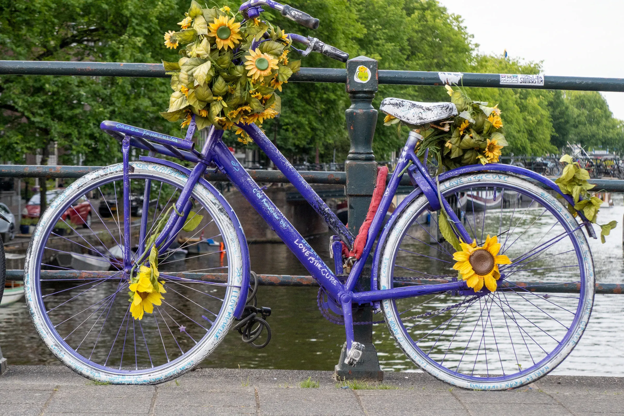 A vivid purple bike decorated with sunflowers, locked to a canal bridge railing, canal behind