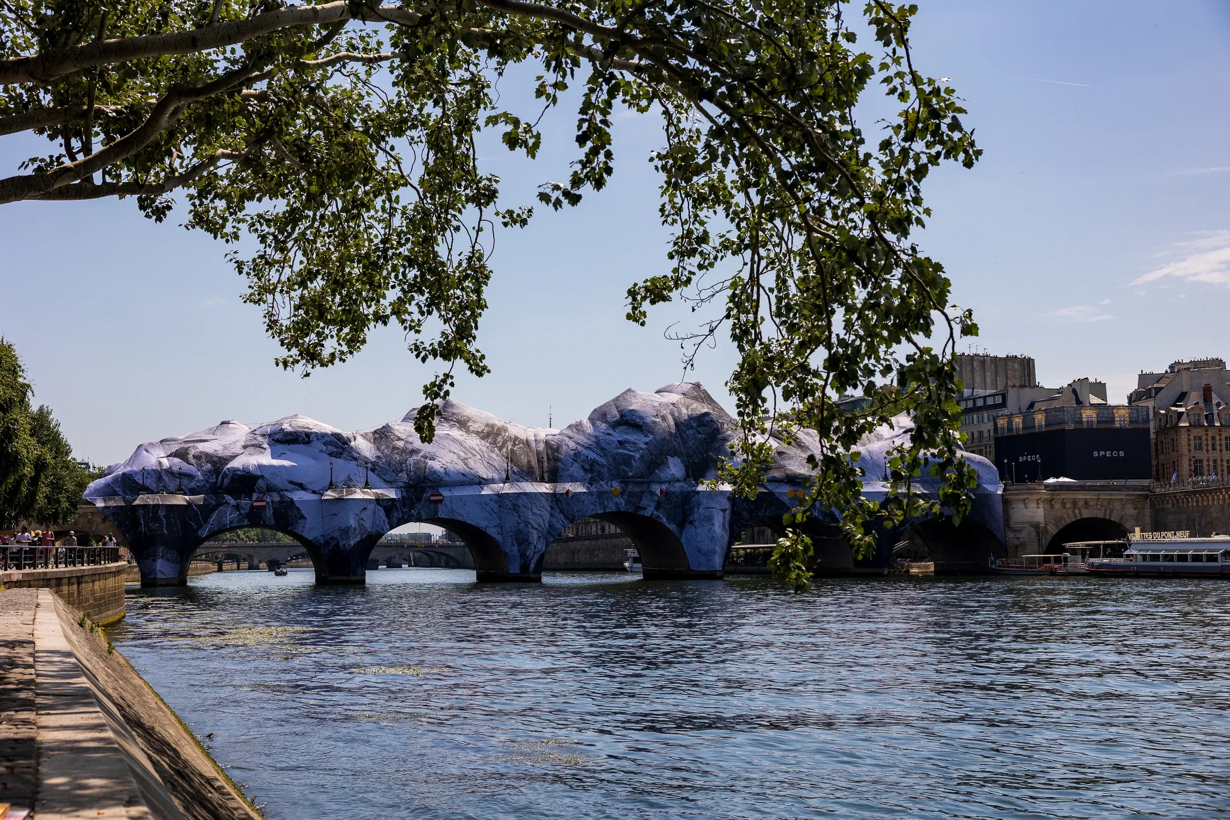 Overhanging tree branches framing a wide view of the Pont Neuf wrapped in grey and white trompe-l'œil rock panels, Paris