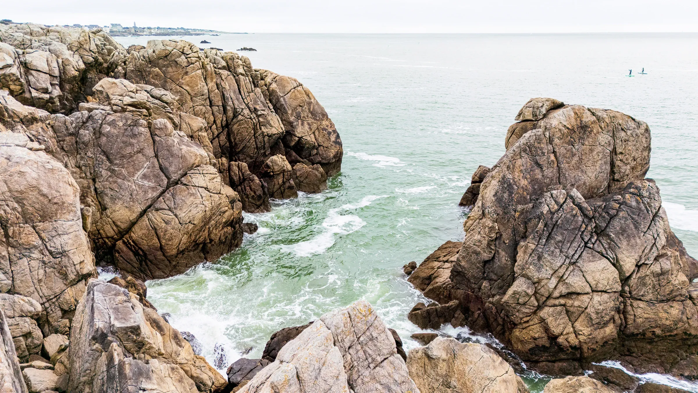 Large split granite boulders at the shoreline, green-tinged sea water, two stand-up paddleboarders in the distance