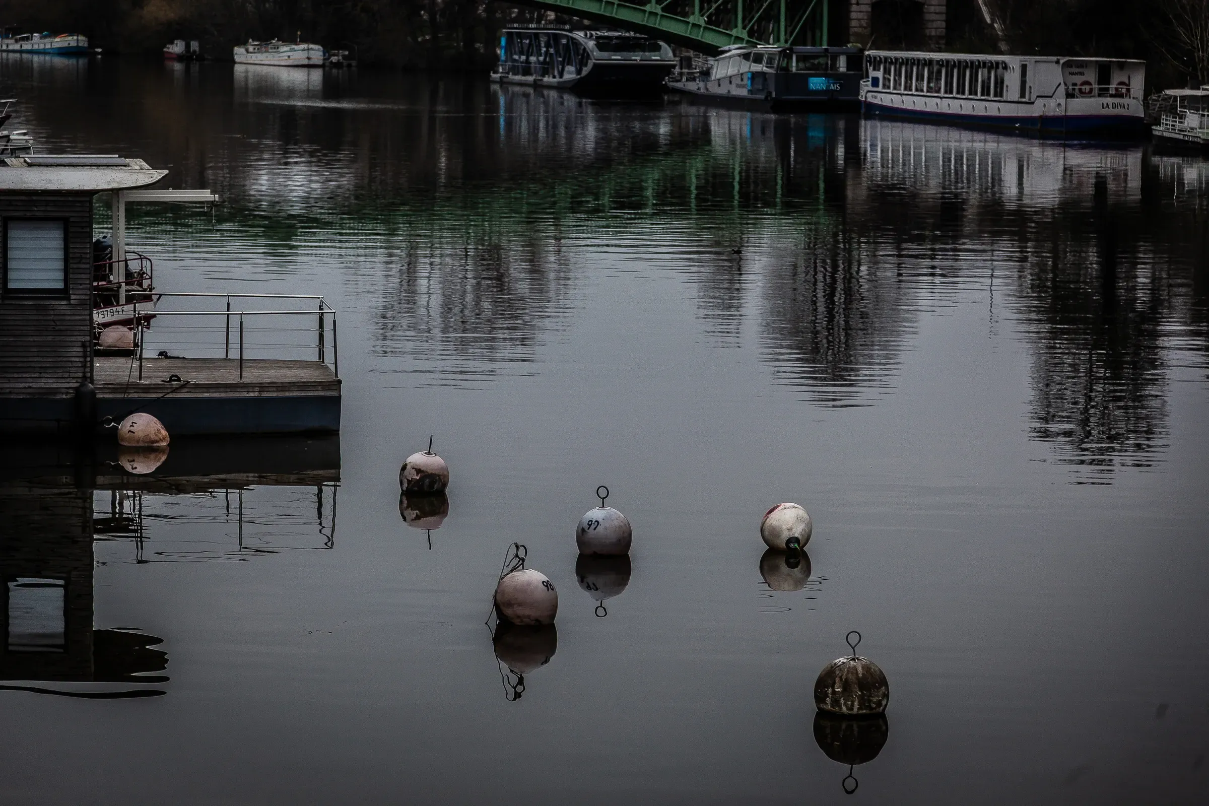 Five white mooring buoys floating on a mirror-still dark river, houseboats and a green bridge reflected behind