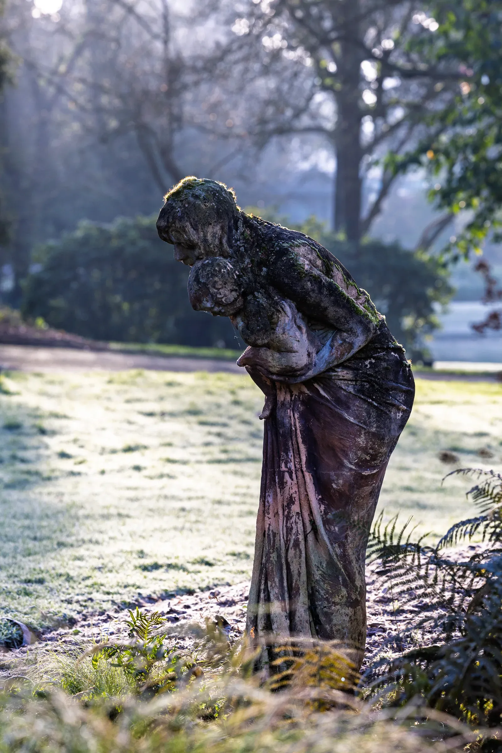 Weathered stone statue of a woman clutching a child, both hunching forward, frosted grass and hazy winter light, Nantes