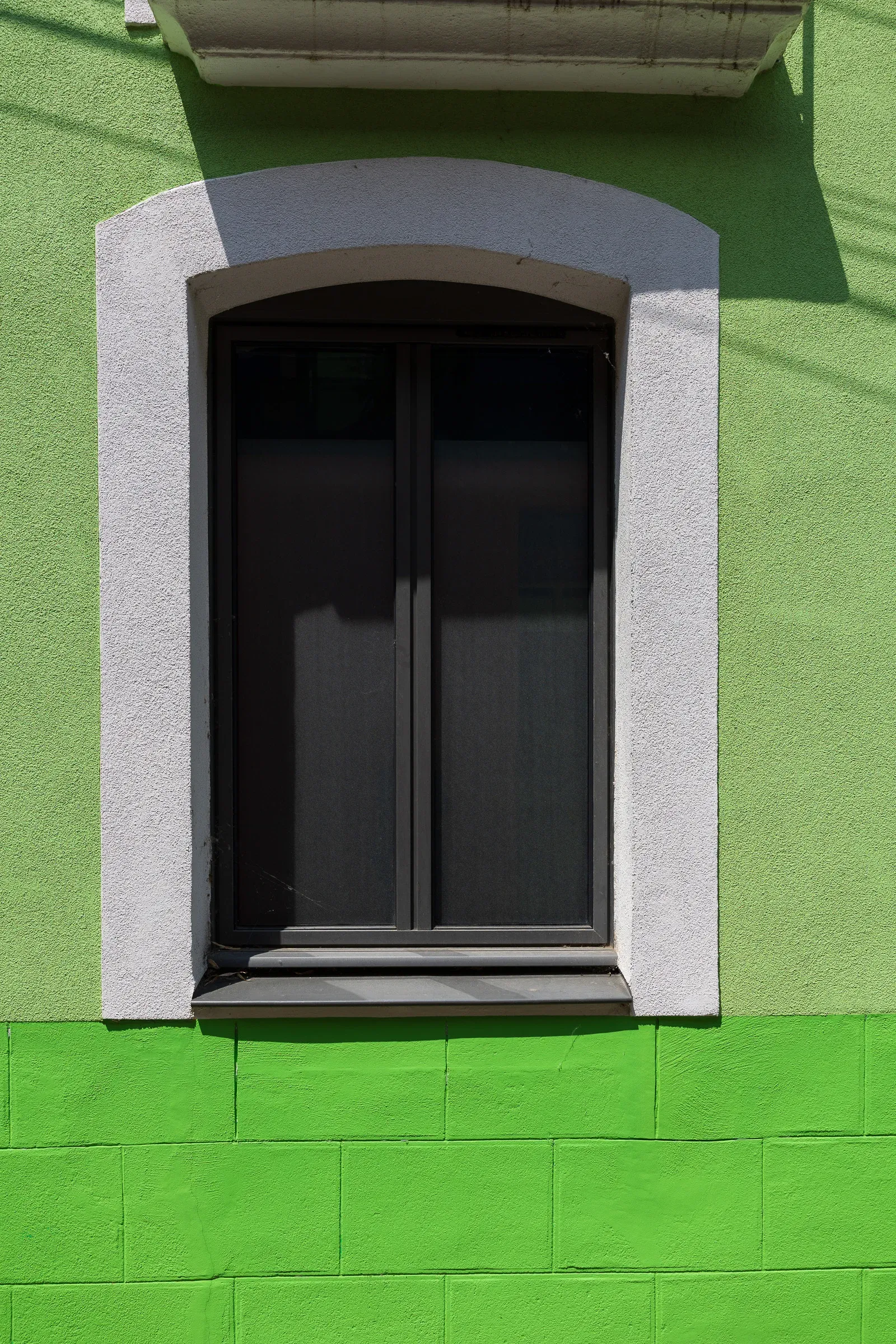 A dark arched window with a grey stone surround set in a bright green rendered wall, plant shadow cast across it