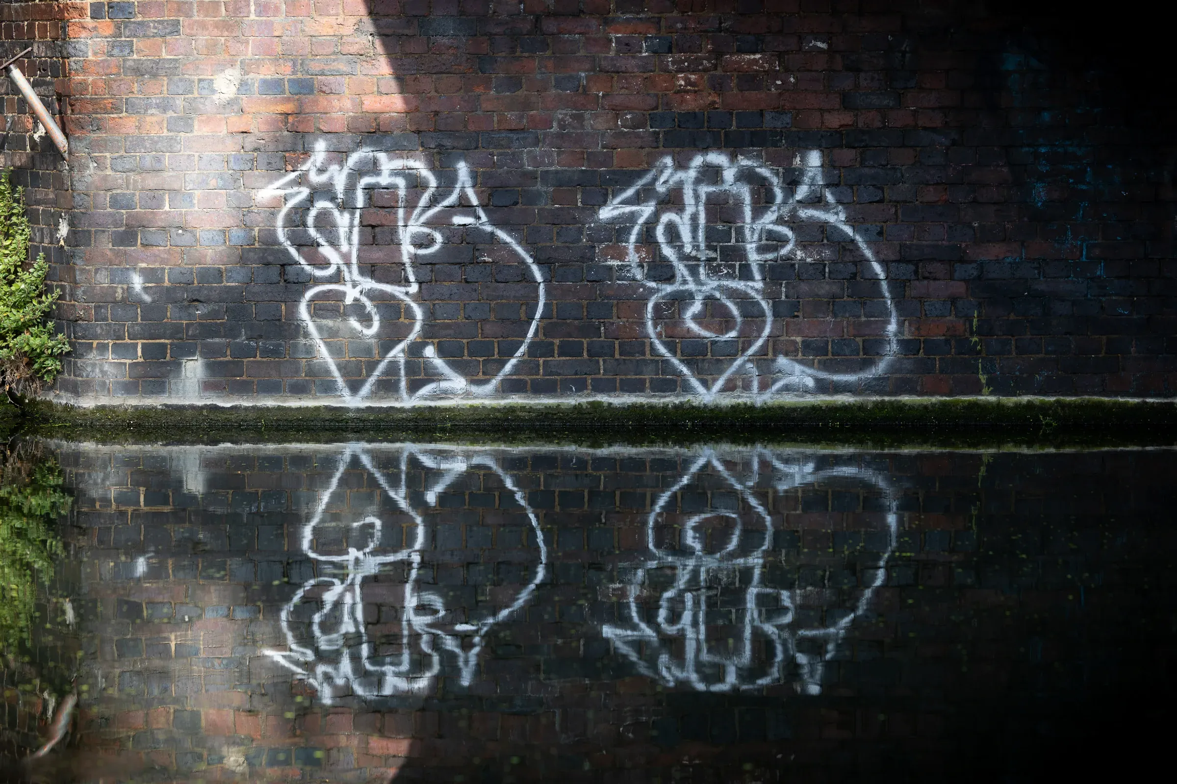 White graffiti tags on a dark brick tunnel wall under a canal bridge, reflected in the still water below