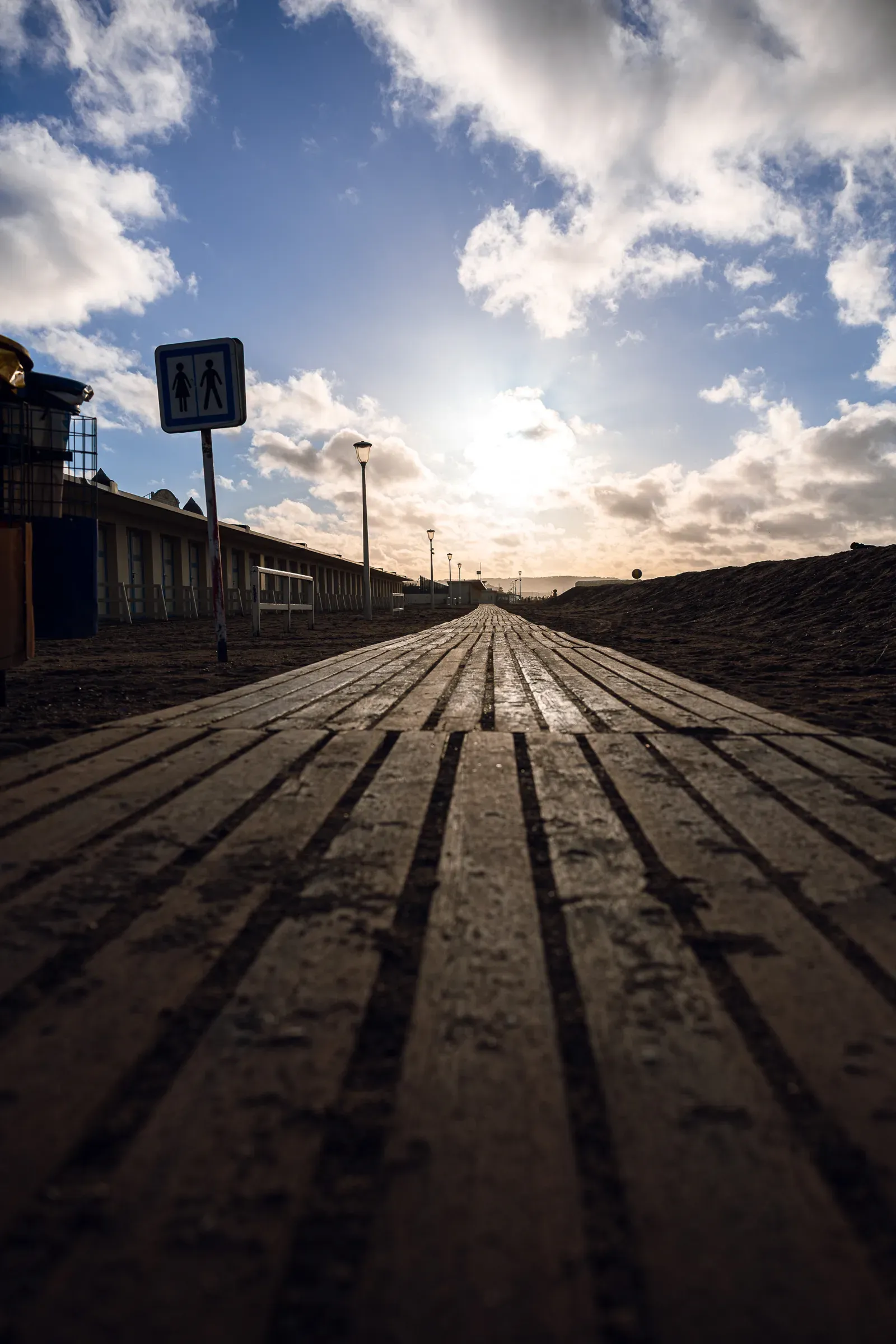 Low-angle shot along a wet wooden boardwalk stretching toward the horizon at low sun, pedestrian sign to the left, beach huts alongside, Trouville