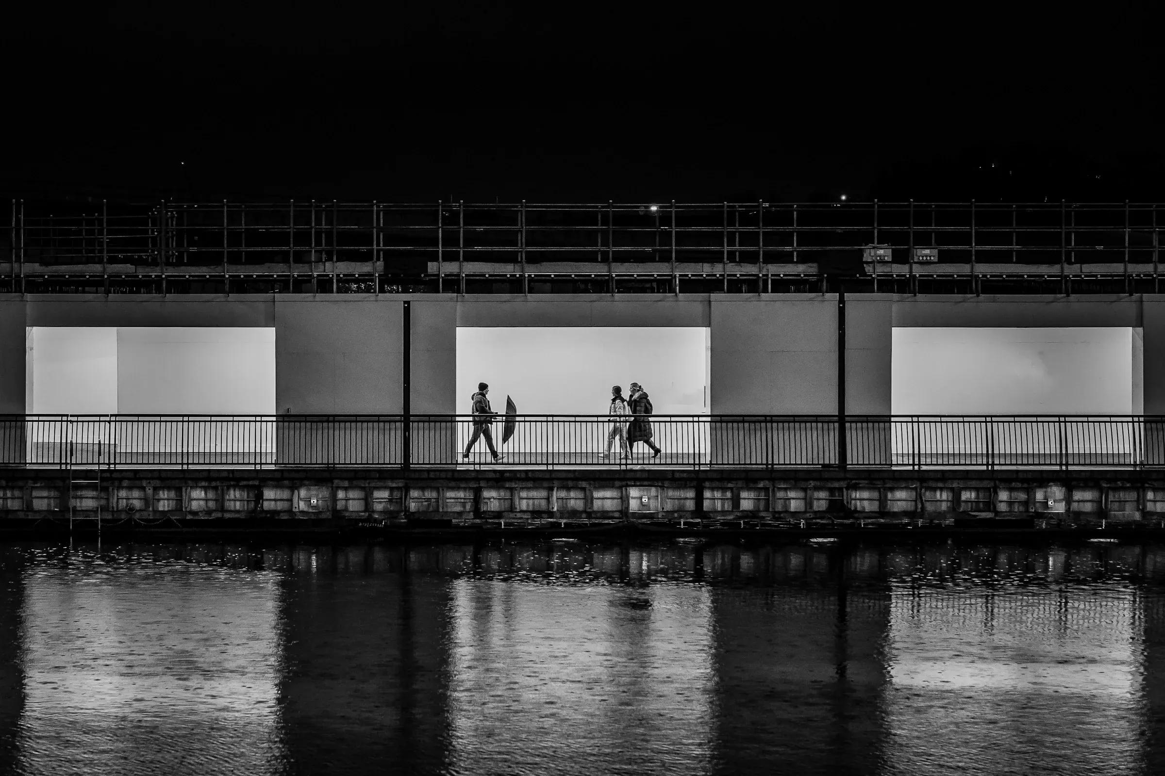 Three people walking along a riverside walkway at night, past illuminated white panels, reflected in the water below