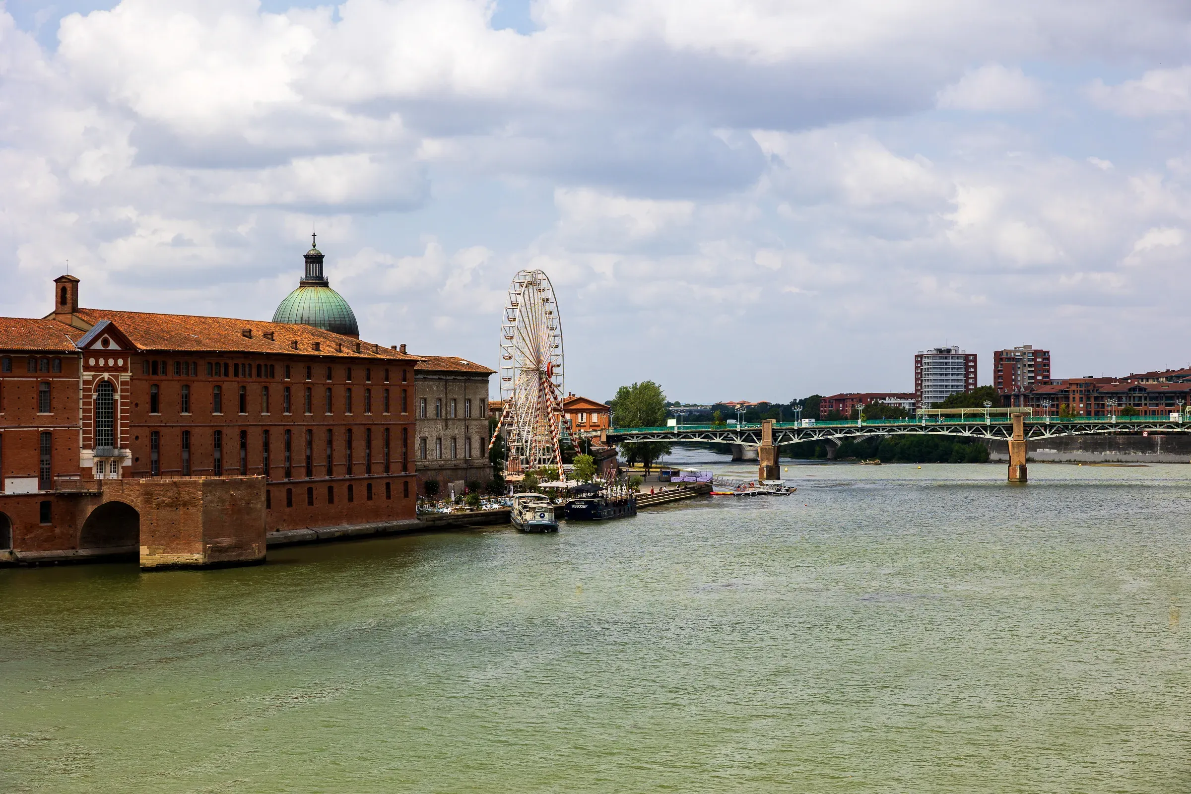 The Garonne river with the Hôtel-Dieu Saint-Jacques brick building and green dome on the left, a Ferris wheel and green iron bridge in the background, Toulouse