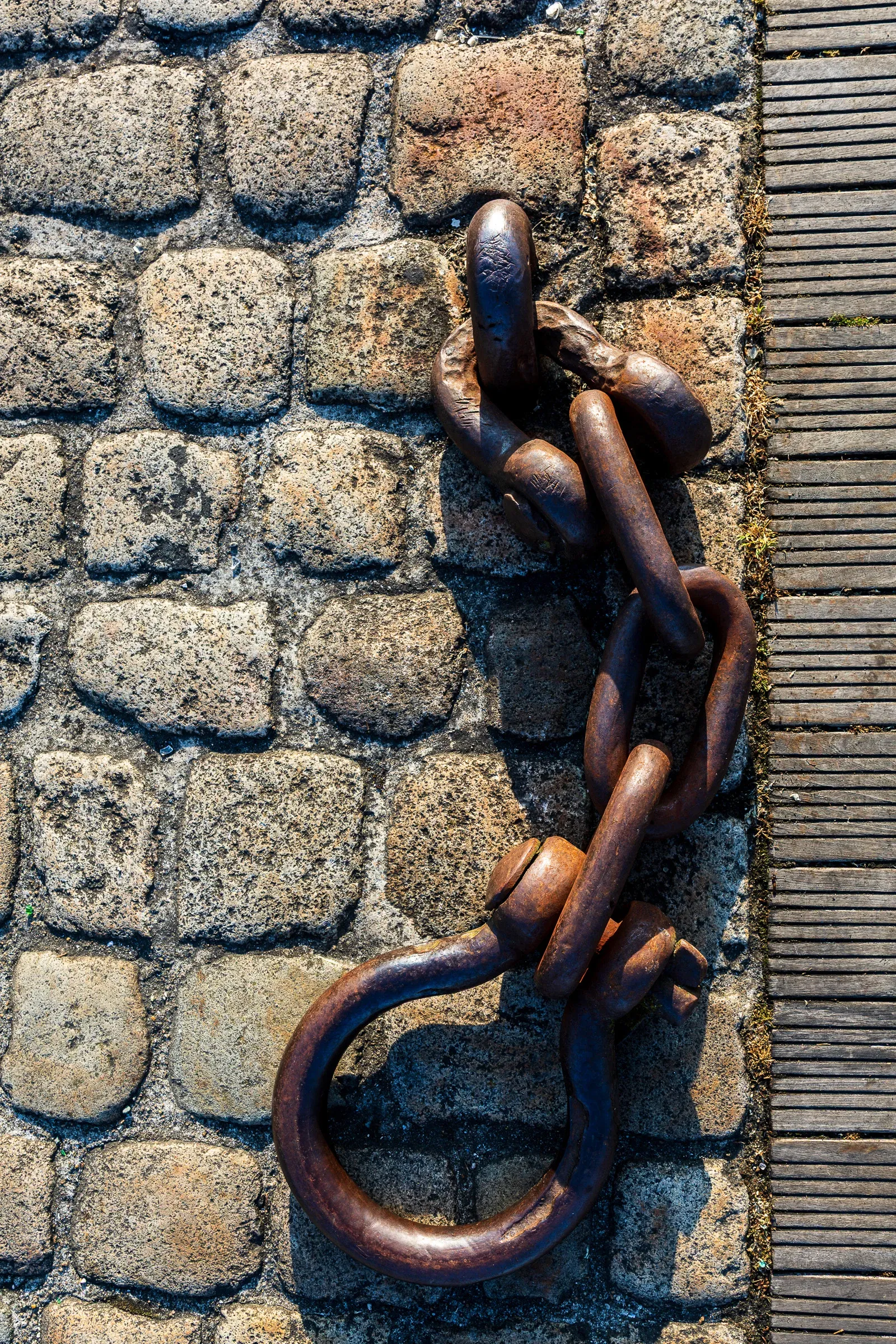 Close-up of a rusted iron mooring ring and knotted chain on granite cobblestones, shot from above in bright direct light, wooden dock edge to the right