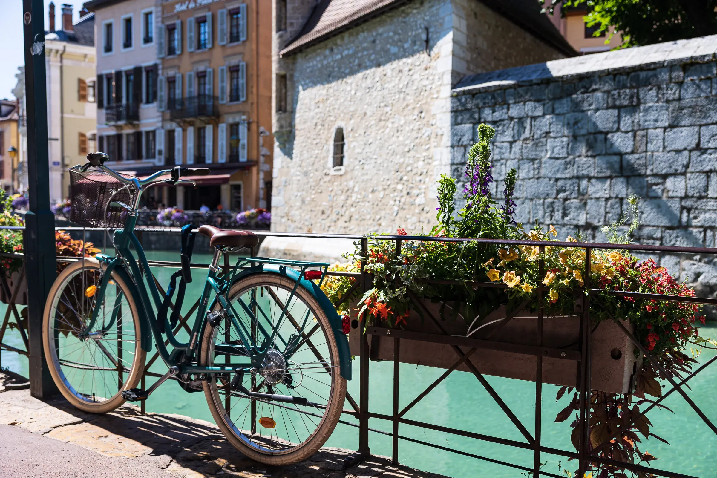 Teal city bicycle locked to a flower-box railing on a bridge, Palais de l'Isle and old town behind, Annecy