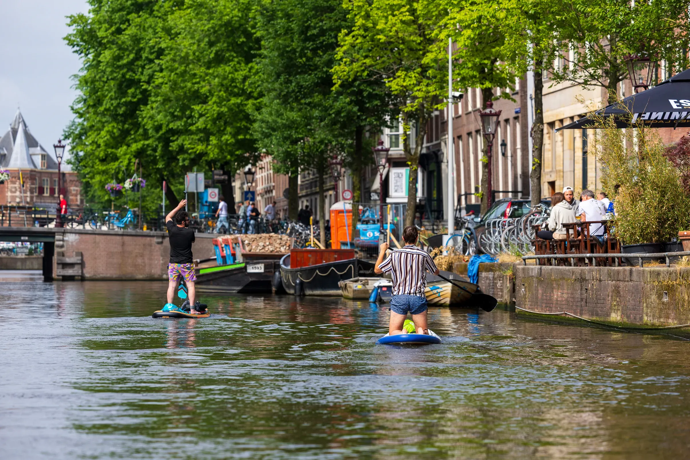 Two people stand-up paddleboarding on an Amsterdam canal, terrace diners visible on the right bank