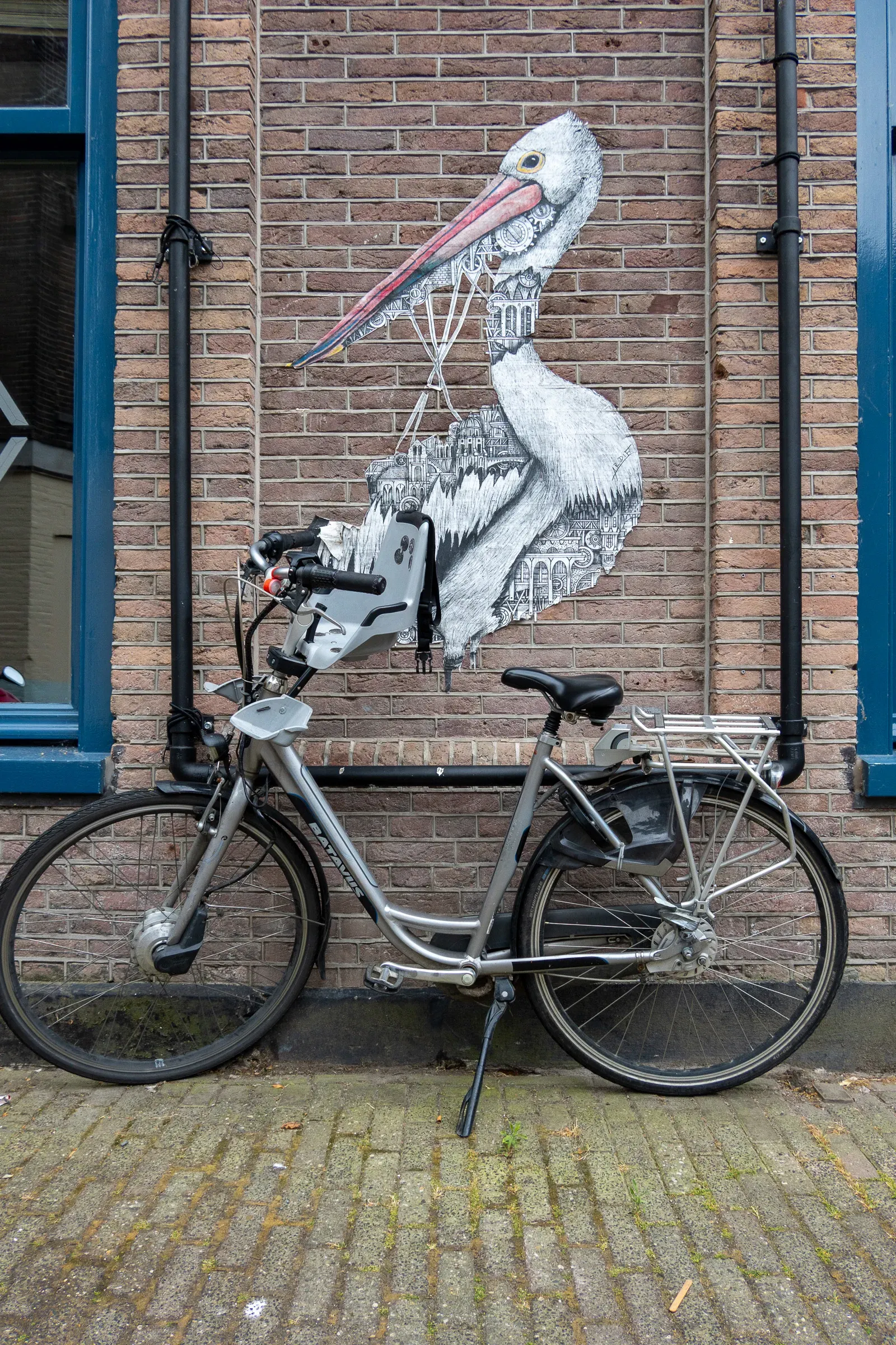 A silver cargo bike parked against a brick wall with a large pelican mural painted on it, Amsterdam