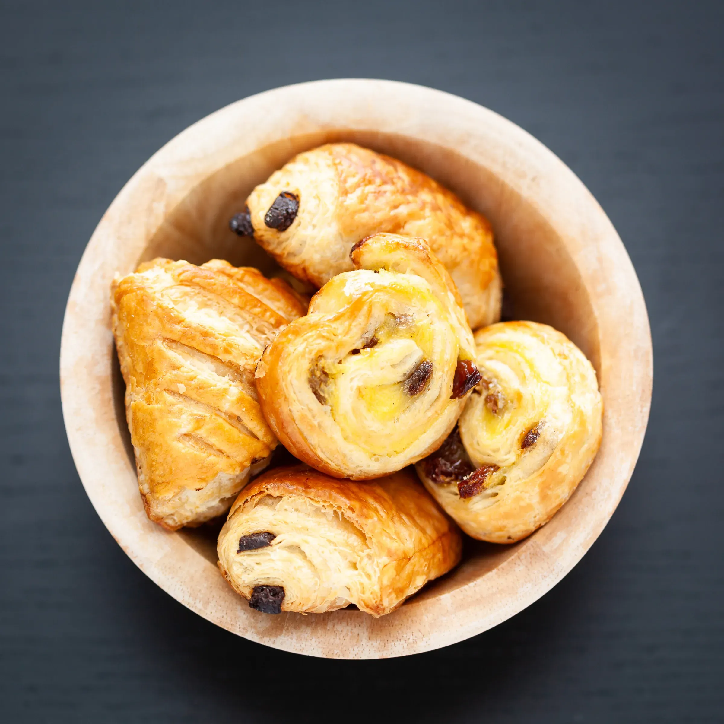 Overhead view of a paper-lined wooden bowl filled with mini croissants and pains aux raisins, dark slate surface beneath