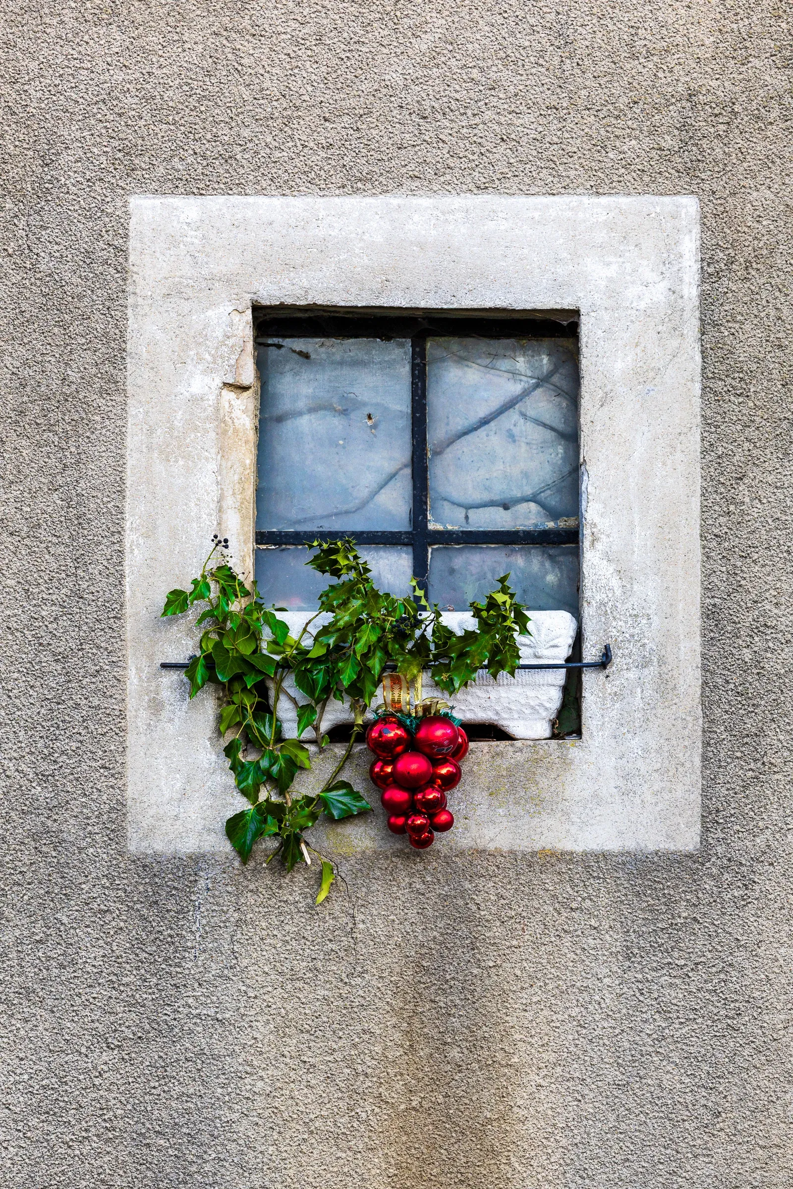 Small stone window on a rough render wall, a sprig of ivy and a cluster of red Christmas ball ornaments hanging below