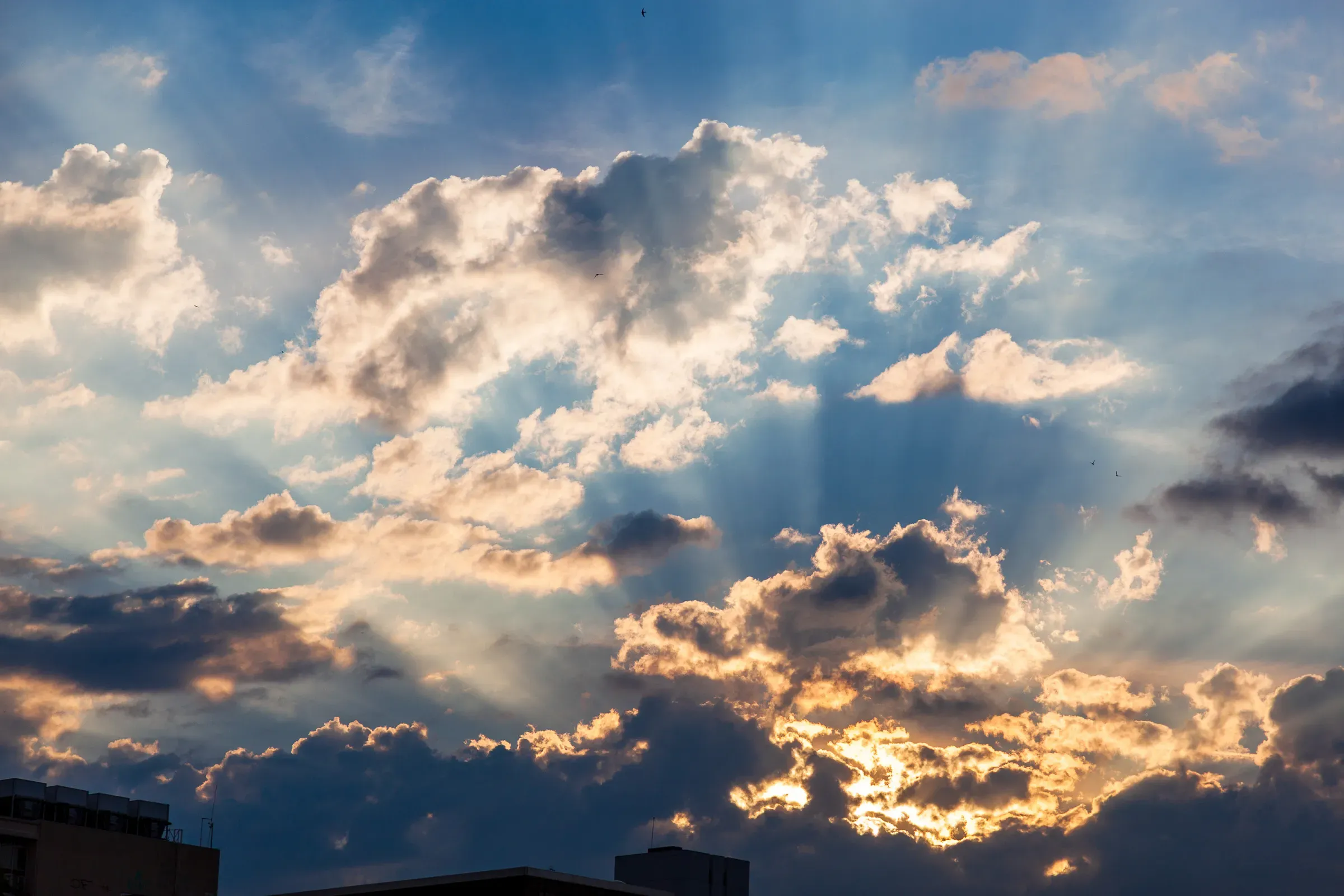 Sun rays breaking through storm clouds at dusk — golden light scattered across a dark blue sky, urban rooftops silhouetted below