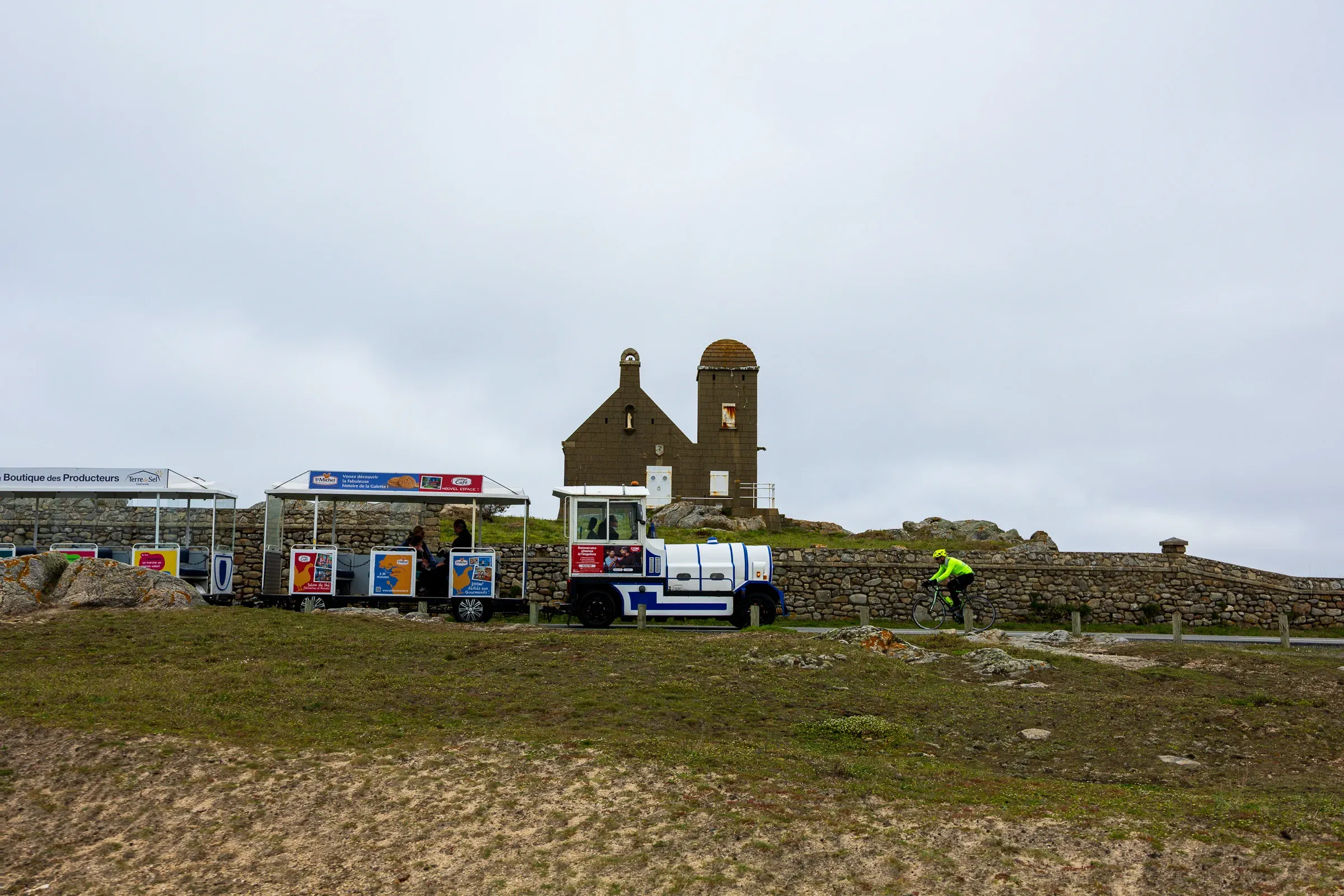 Old stone chapel ruin with twin towers, a seafood market stall with colorful signage in front, a cyclist in yellow passing by