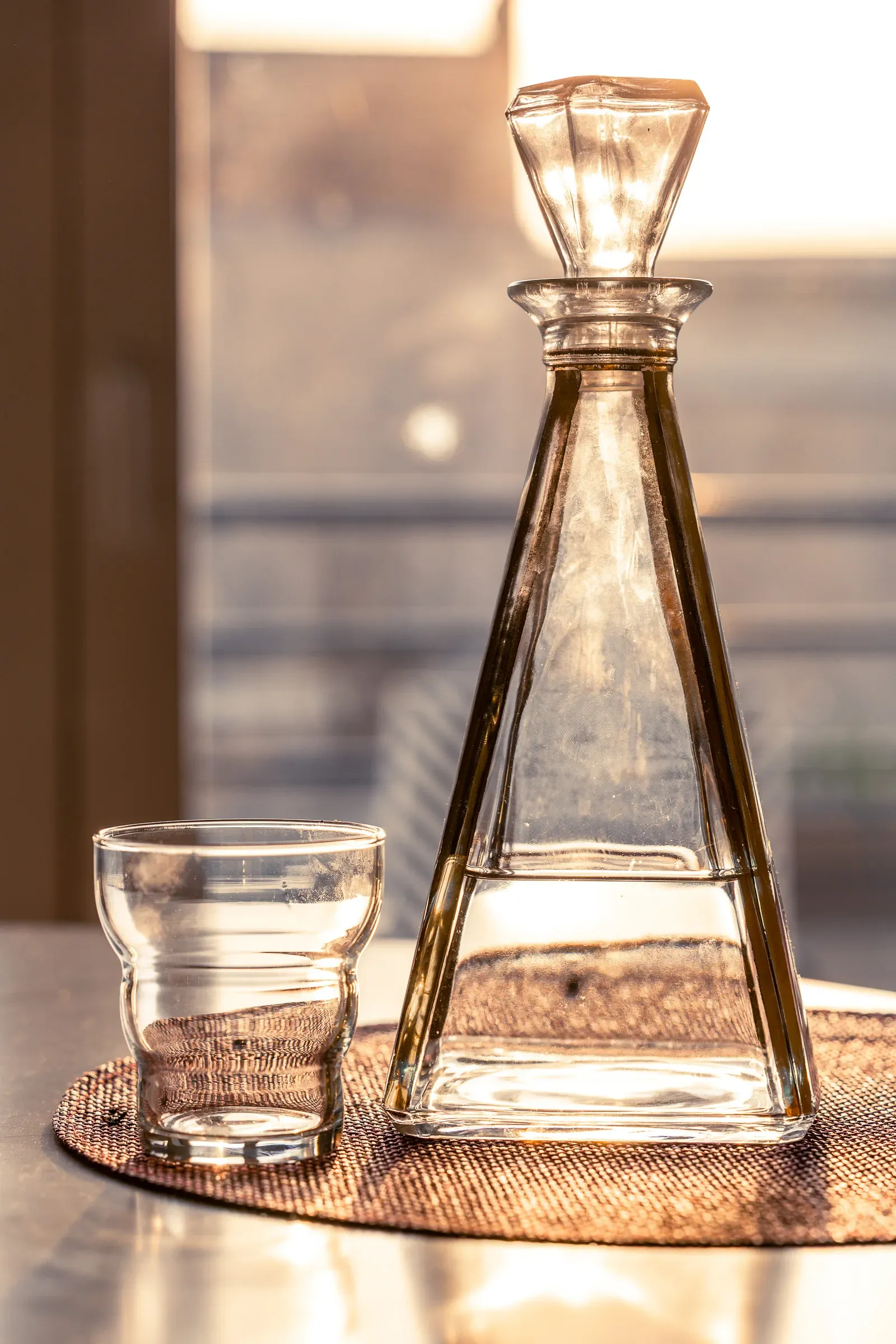 A triangular glass decanter and a small tumbler on a rattan mat, backlit by warm golden afternoon window light