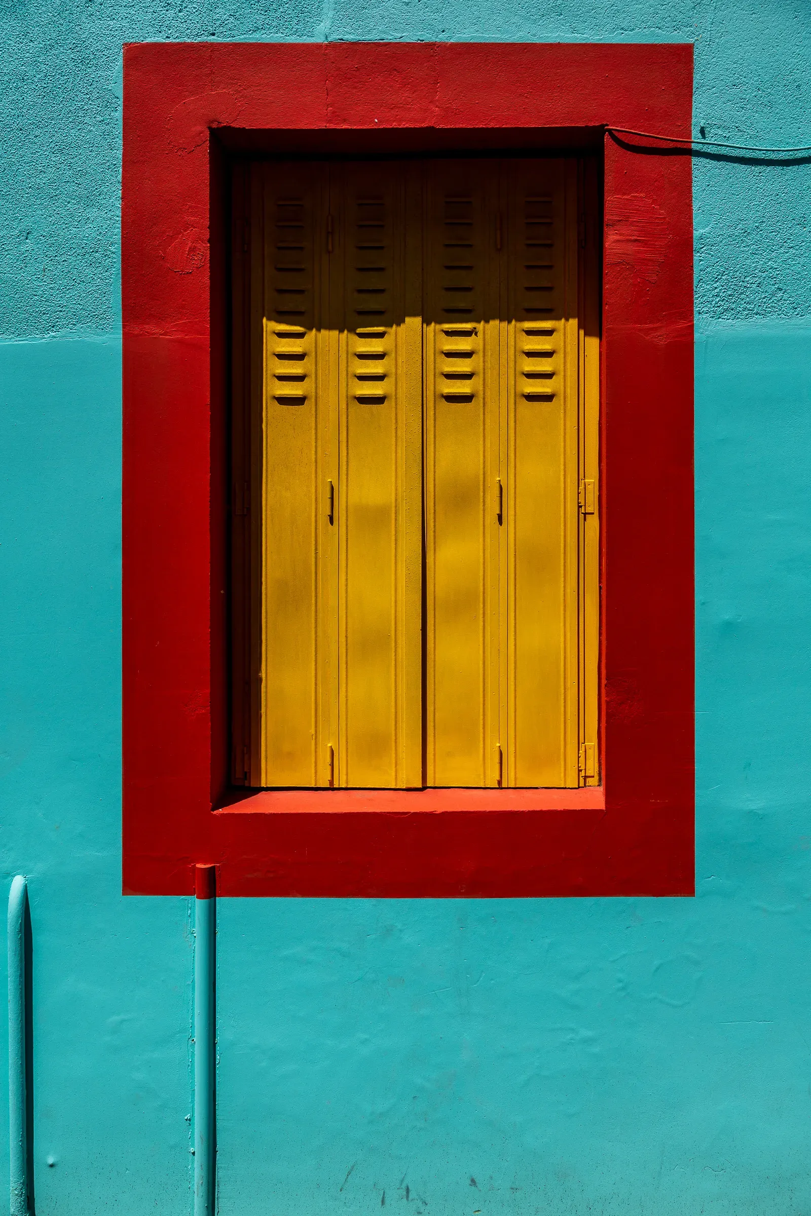 Yellow louvered metal shutters in a red-framed window recess on a turquoise rendered wall, Trentemoult